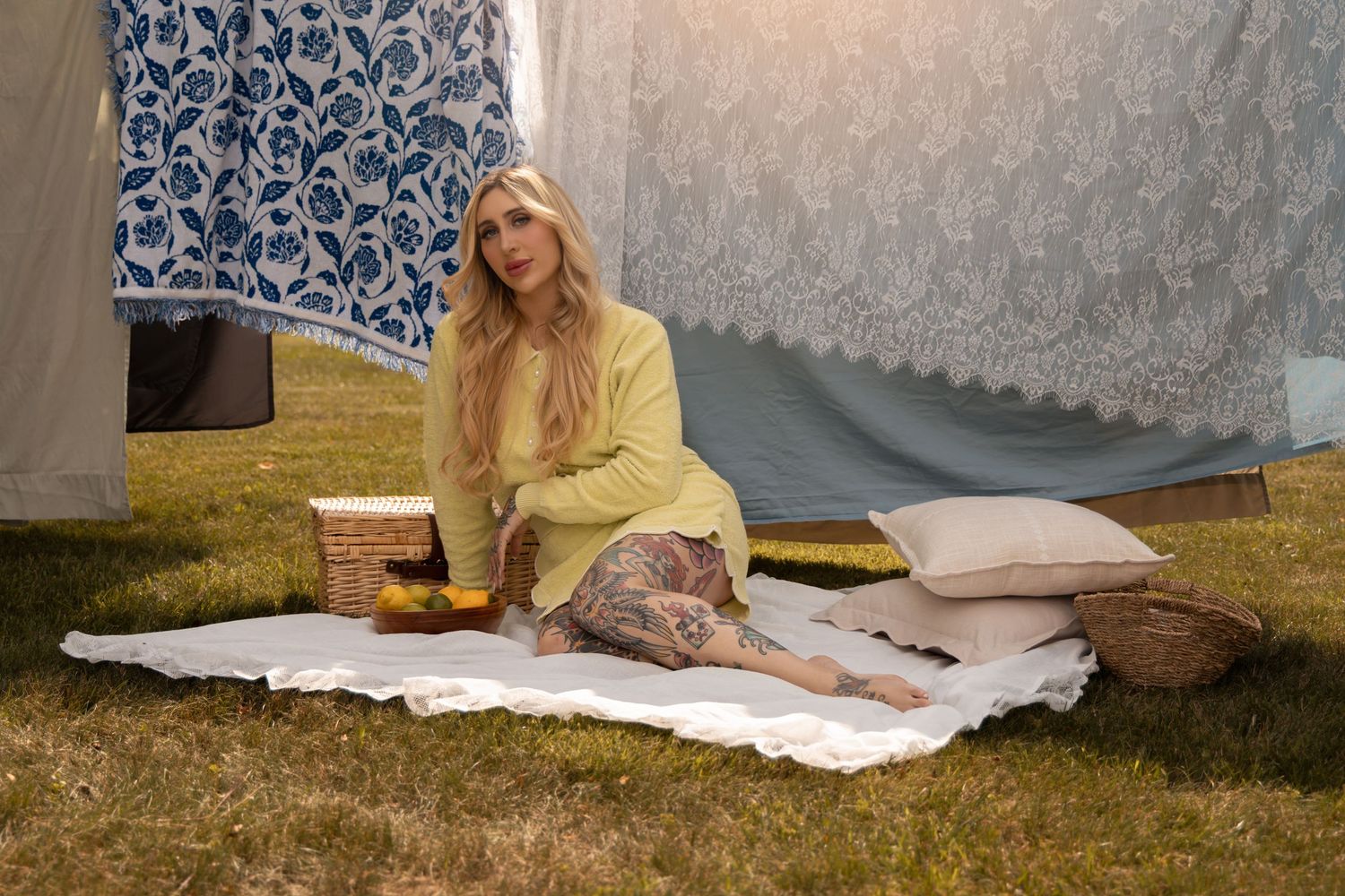 Woman in yellow top and floral skirt sitting on white blanket outdoors with hanging fabrics and wicker basket.