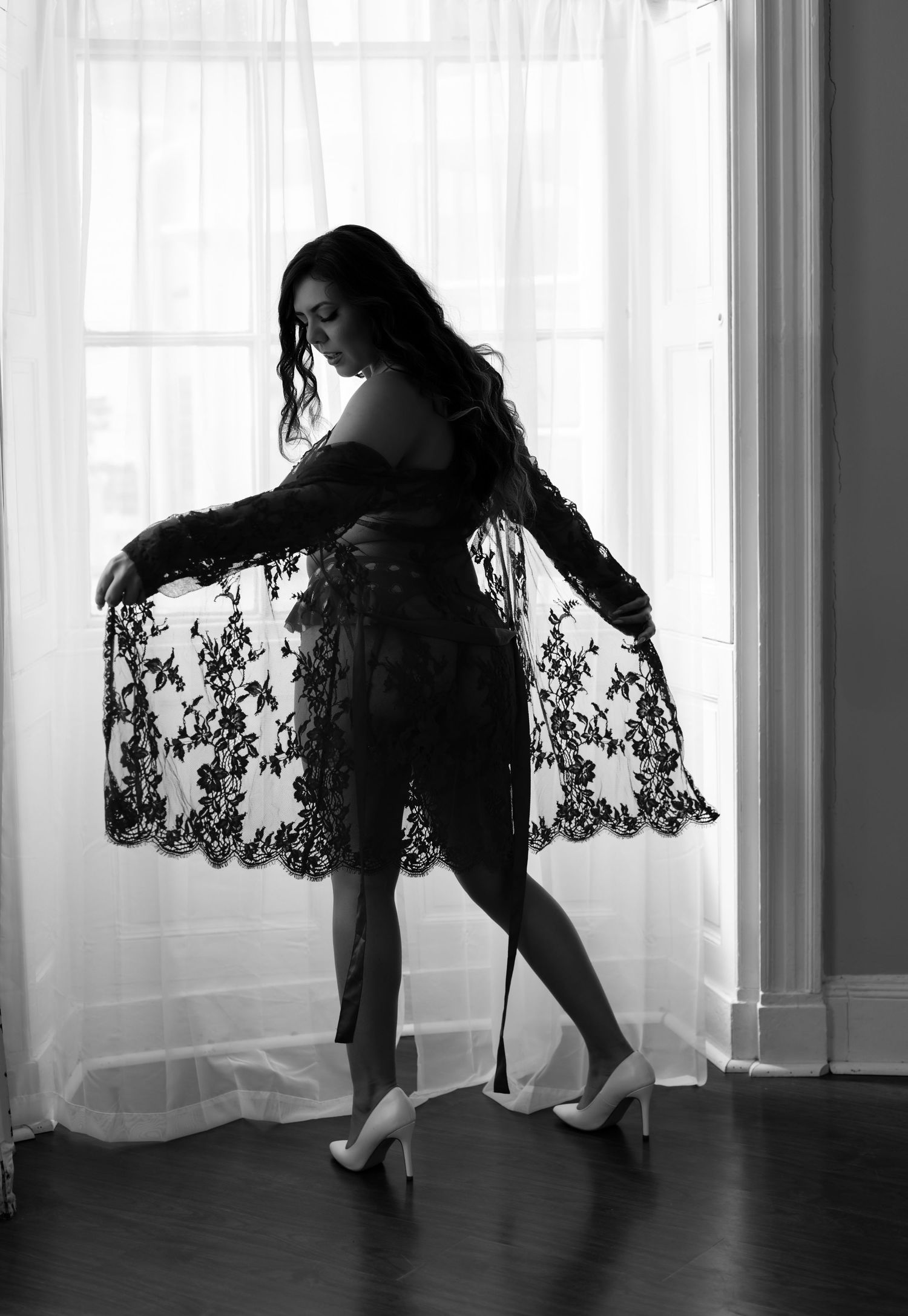 Silhouetted figure in lace dress poses gracefully by window in black and white photo.