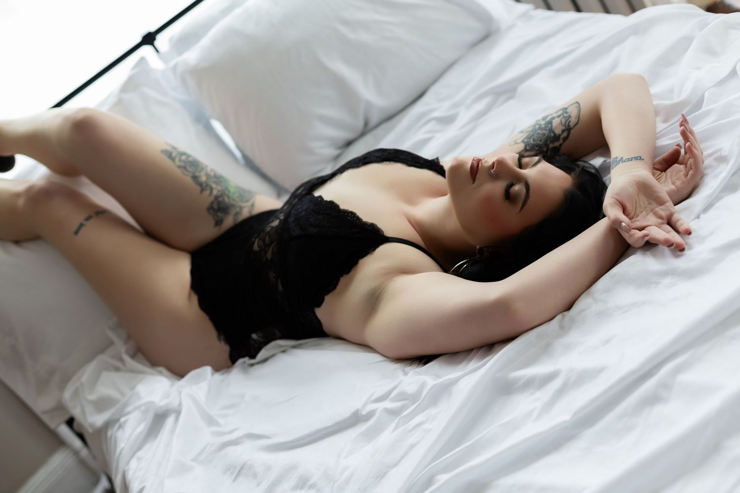 Woman in black lingerie lying on white bedsheets with arms raised above her head.