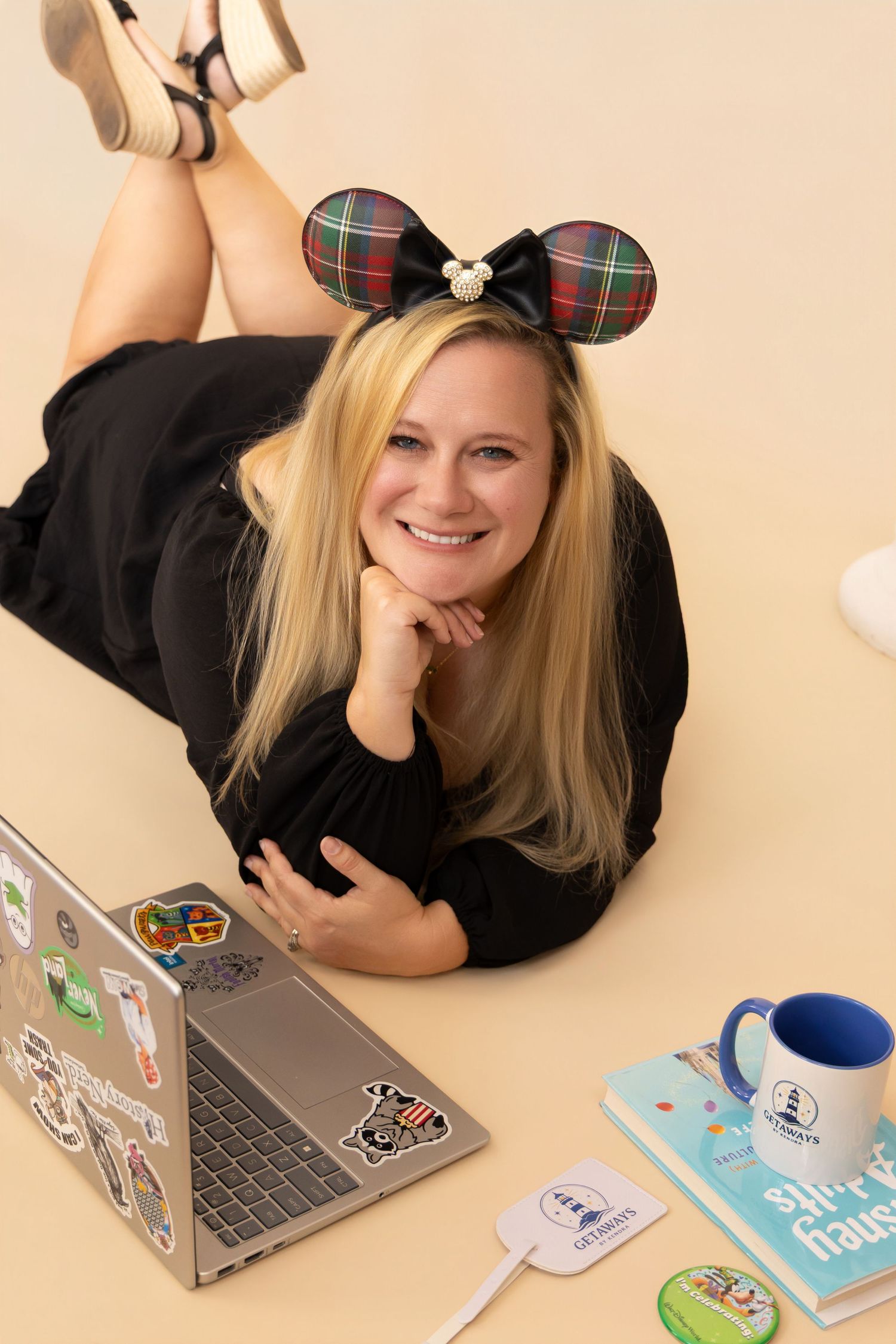 Smiling blonde woman lying on floor with Minnie Mouse ears, laptop with stickers, blue mug, and books on beige background.