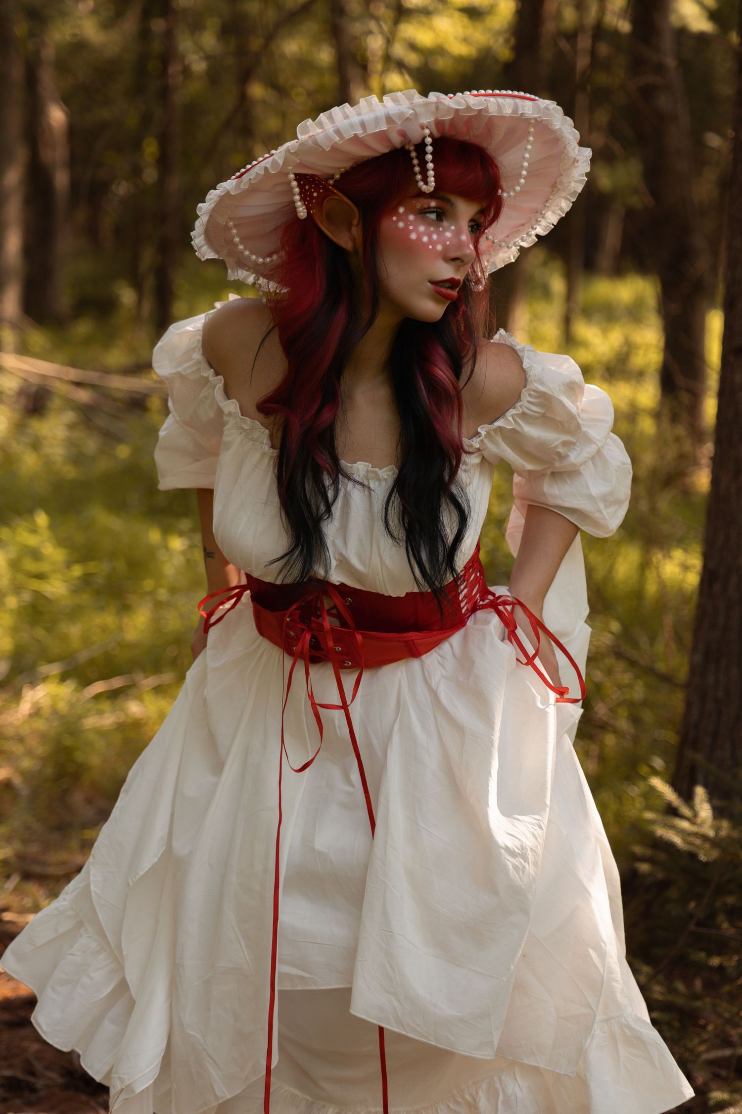 A woman in a white off-shoulder dress with red belt and pearl-adorned hat poses in a forest with fantasy elf cosplay makeup.
