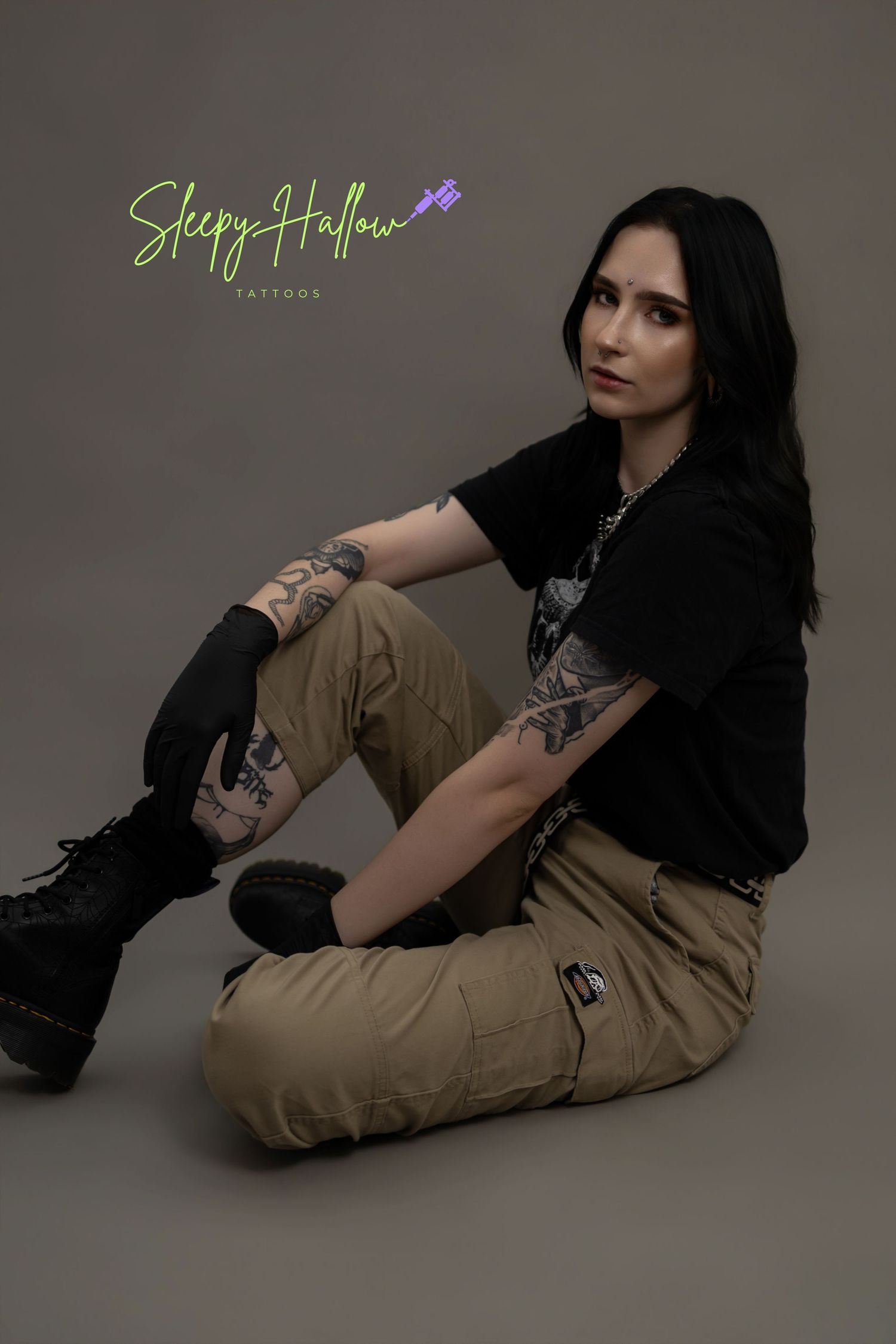 Tattooed person sitting on floor wearing black tee, khaki cargo pants, and boots, posed for Sleepy Hallow Tattoos.