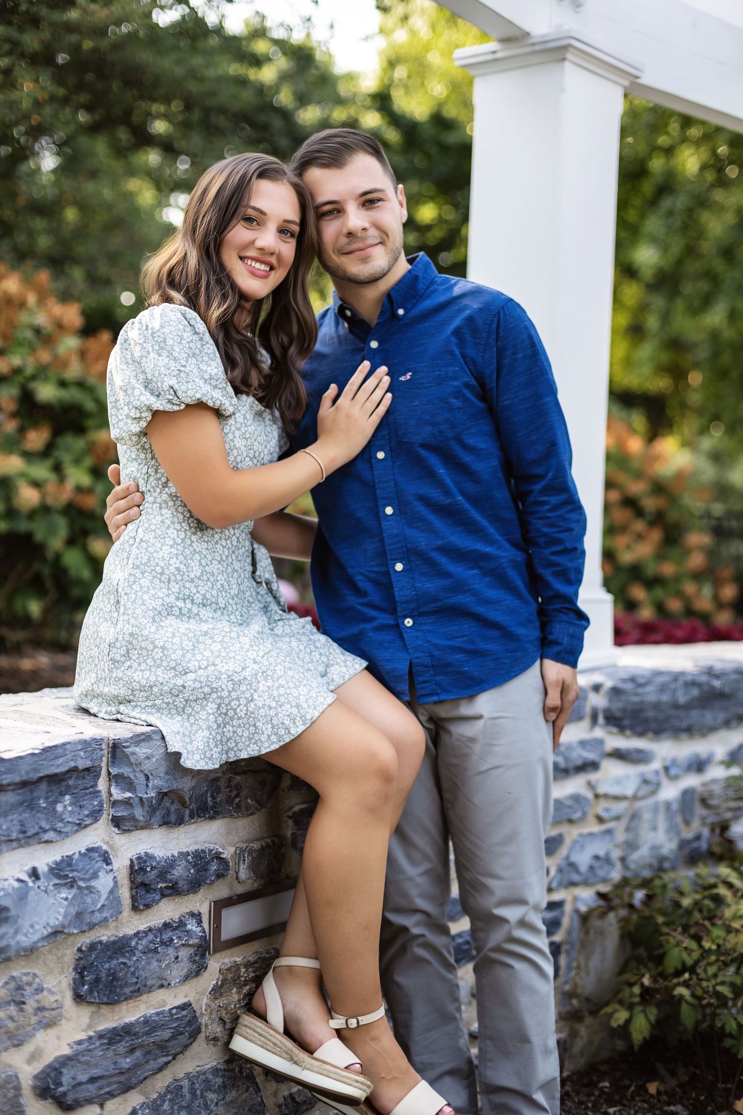 A romantic couple poses together on a stone wall with white columns and greenery in the background.