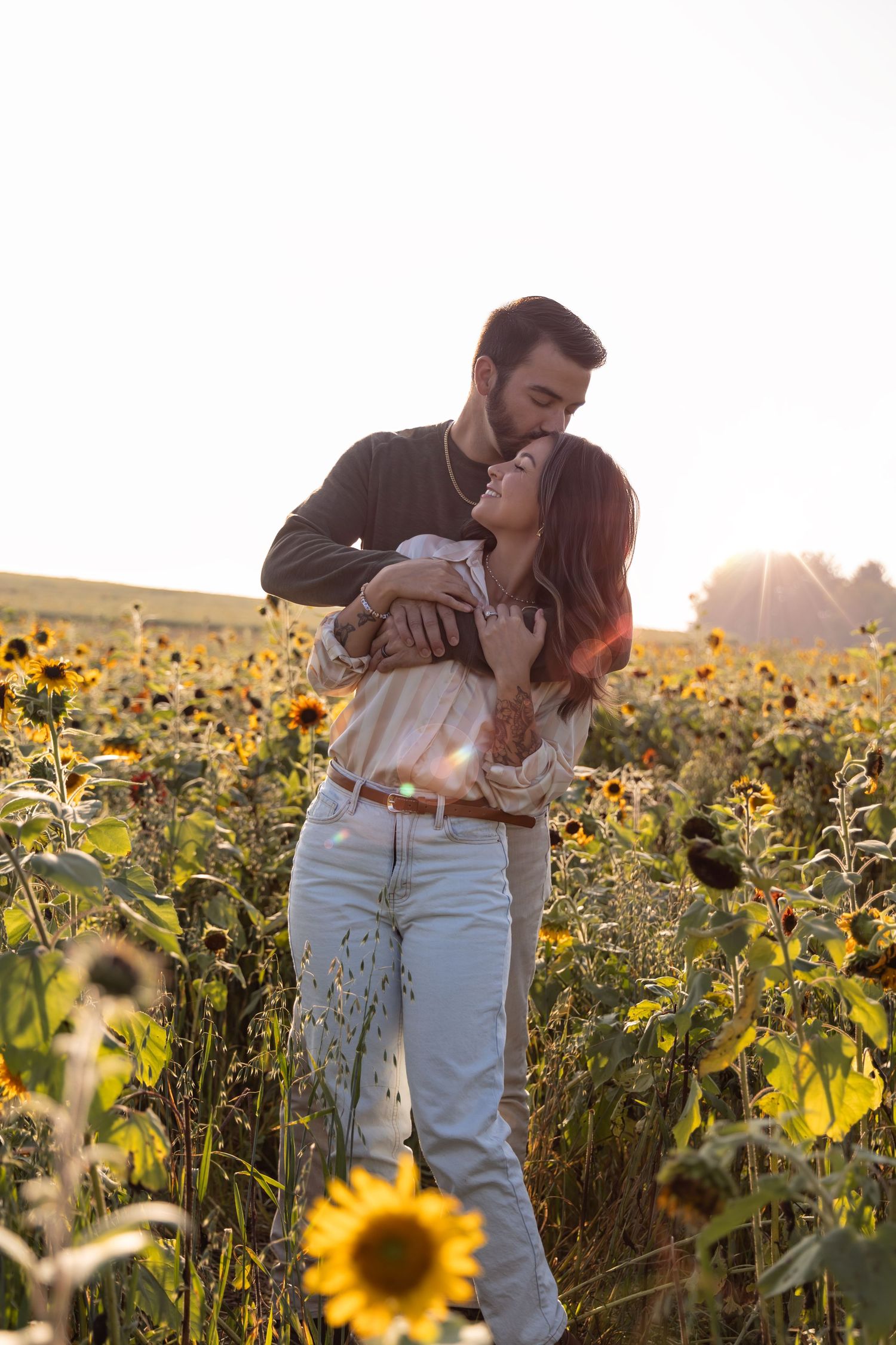 A couple shares romantic moments while standing together in a golden sunflower field at sunset.