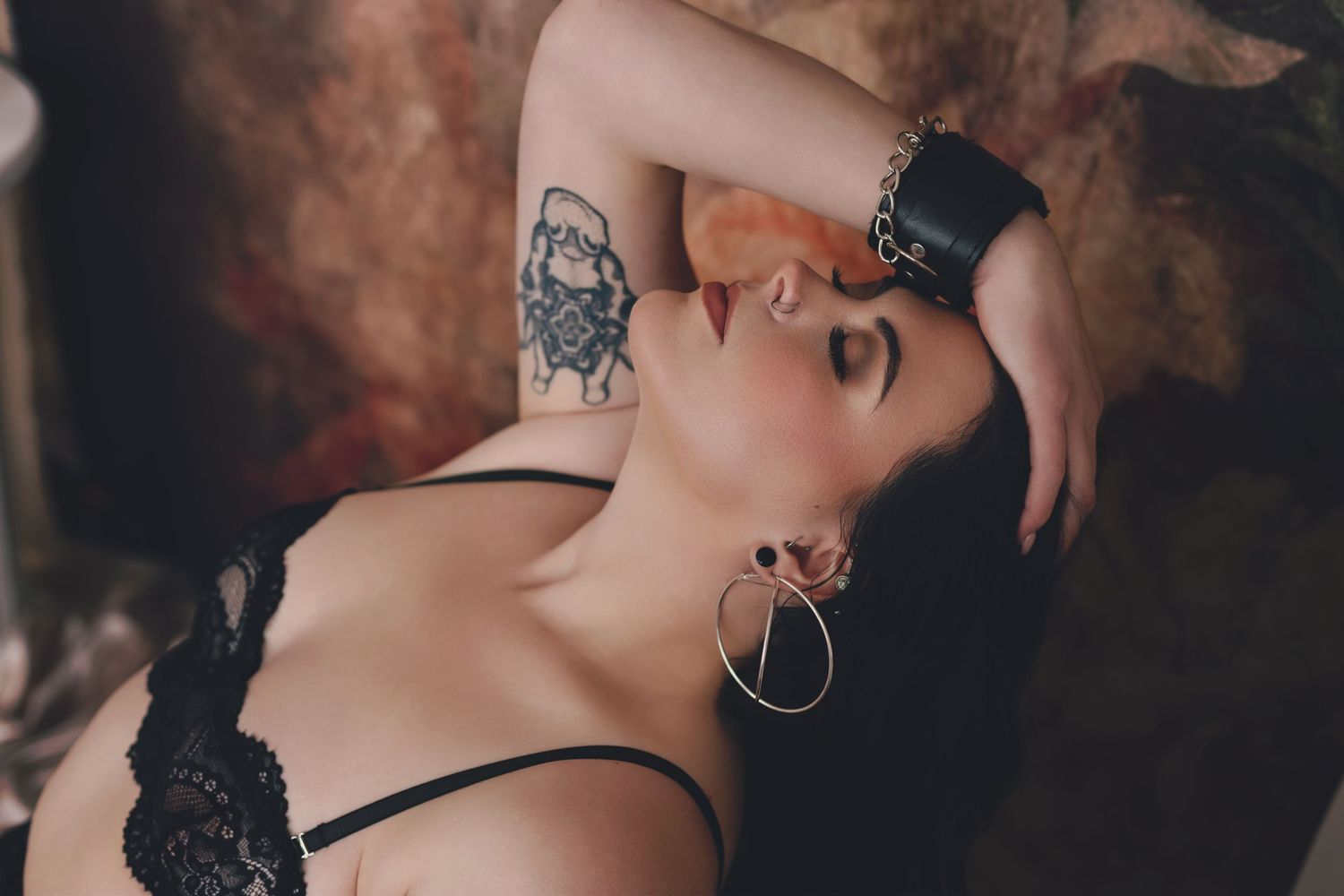 Artistic portrait of someone with a tattoo and hoop earrings posing against dark background in black lingerie.