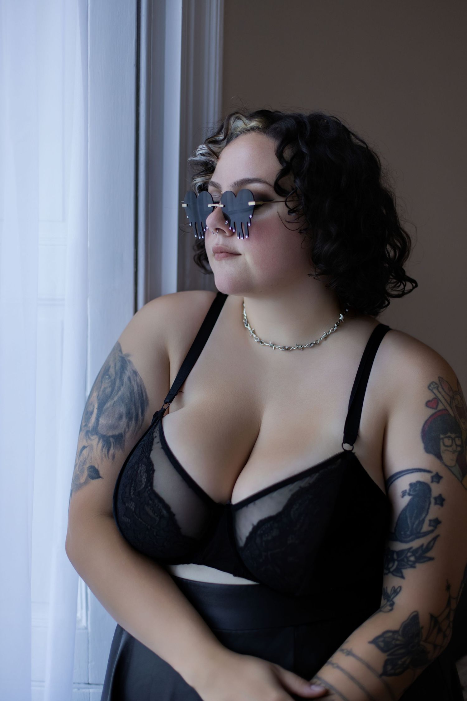 A person wearing black lingerie poses in moody lighting near a window with tattoos visible on their arms.