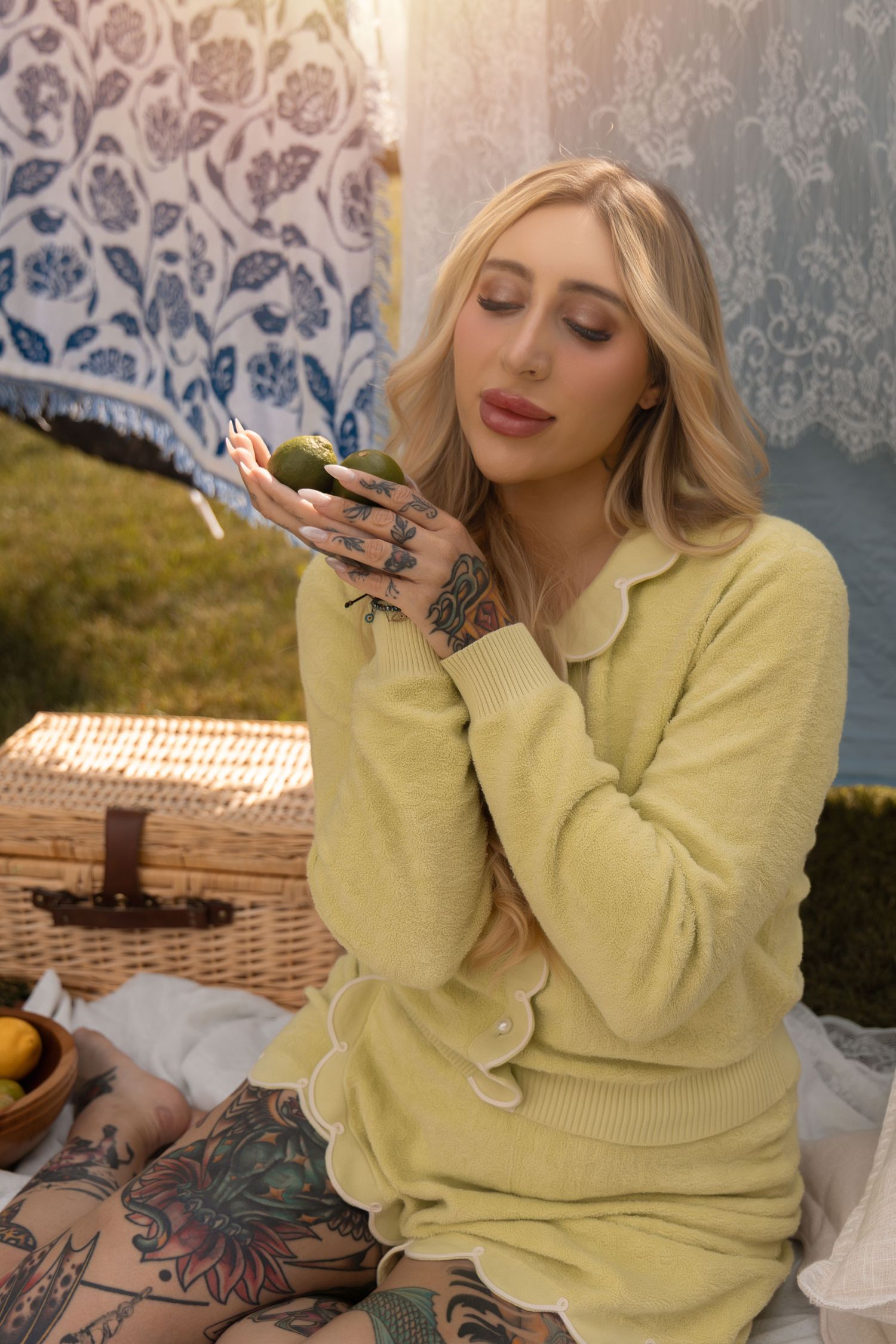 Blonde tattooed woman in yellow sweater holding an avocado at an outdoor picnic with lace curtains.