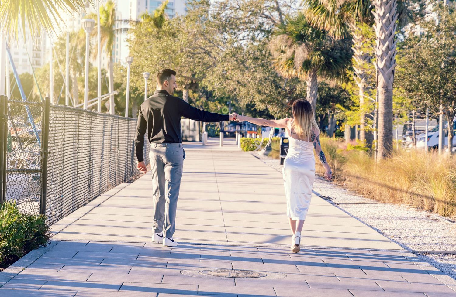 People skating and walking along a sunny waterfront promenade with palm trees and fencing on a clear day.