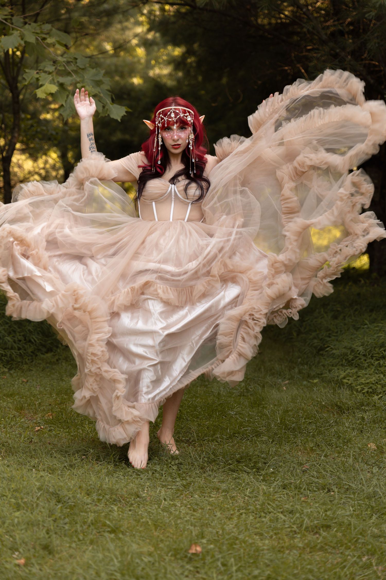 Woman in flowing champagne tulle gown with elf ears and red crown poses outdoors in lush green forest setting.