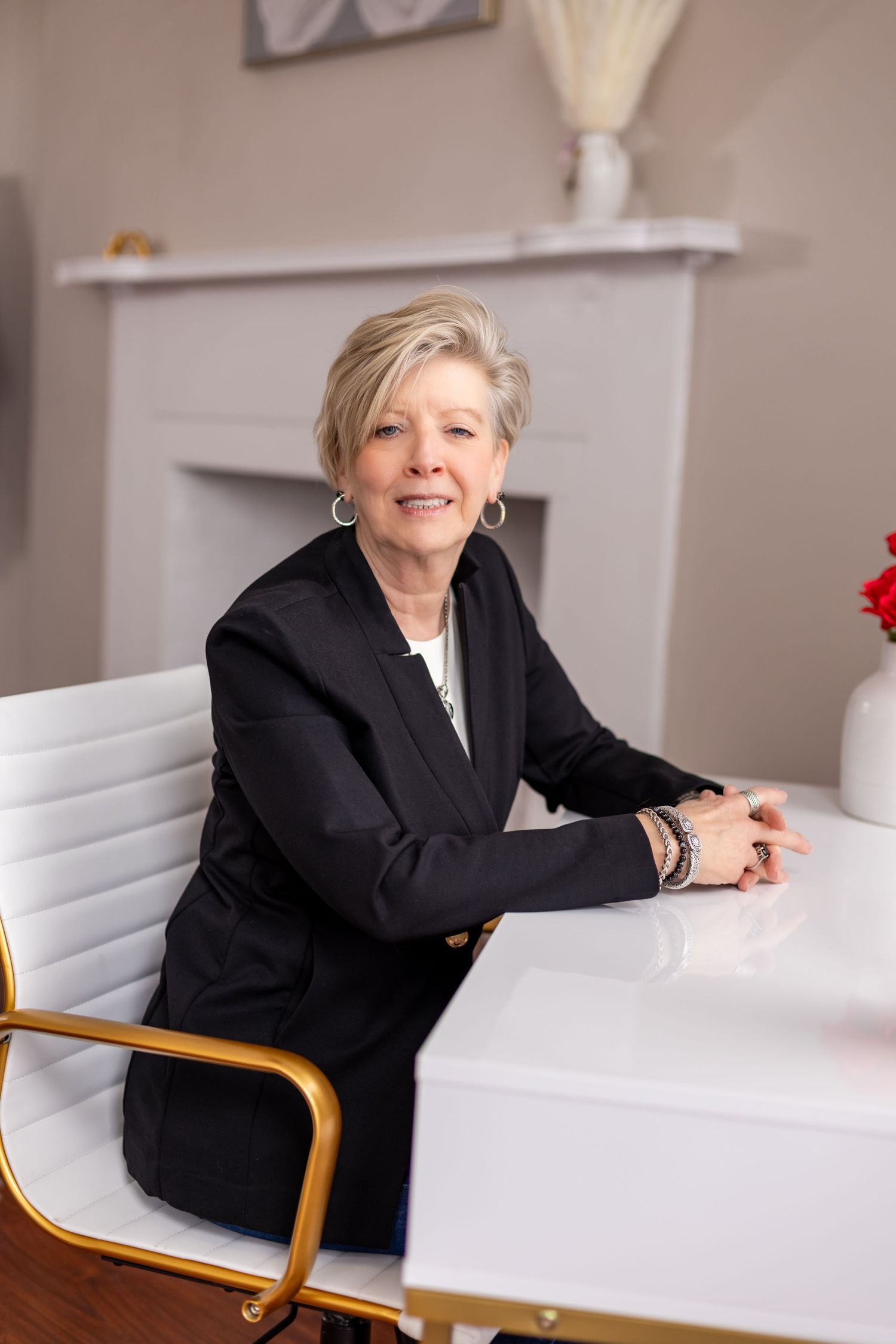 Professional woman in black blazer sits at white desk in elegant office setting.