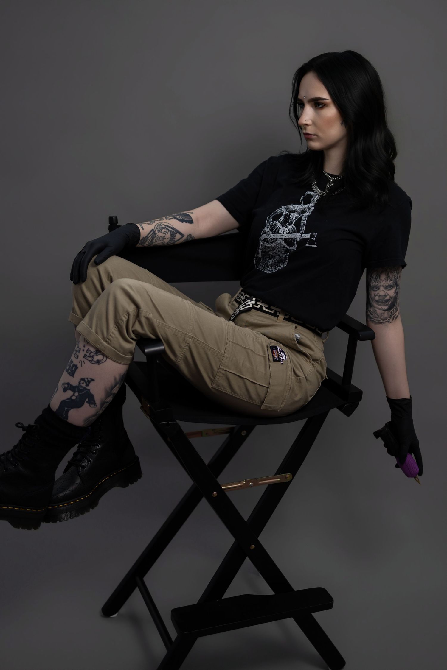 Tattooed female tattoo artist relaxing on a director's chair, wearing black gloves and cargo pants in a studio setting.