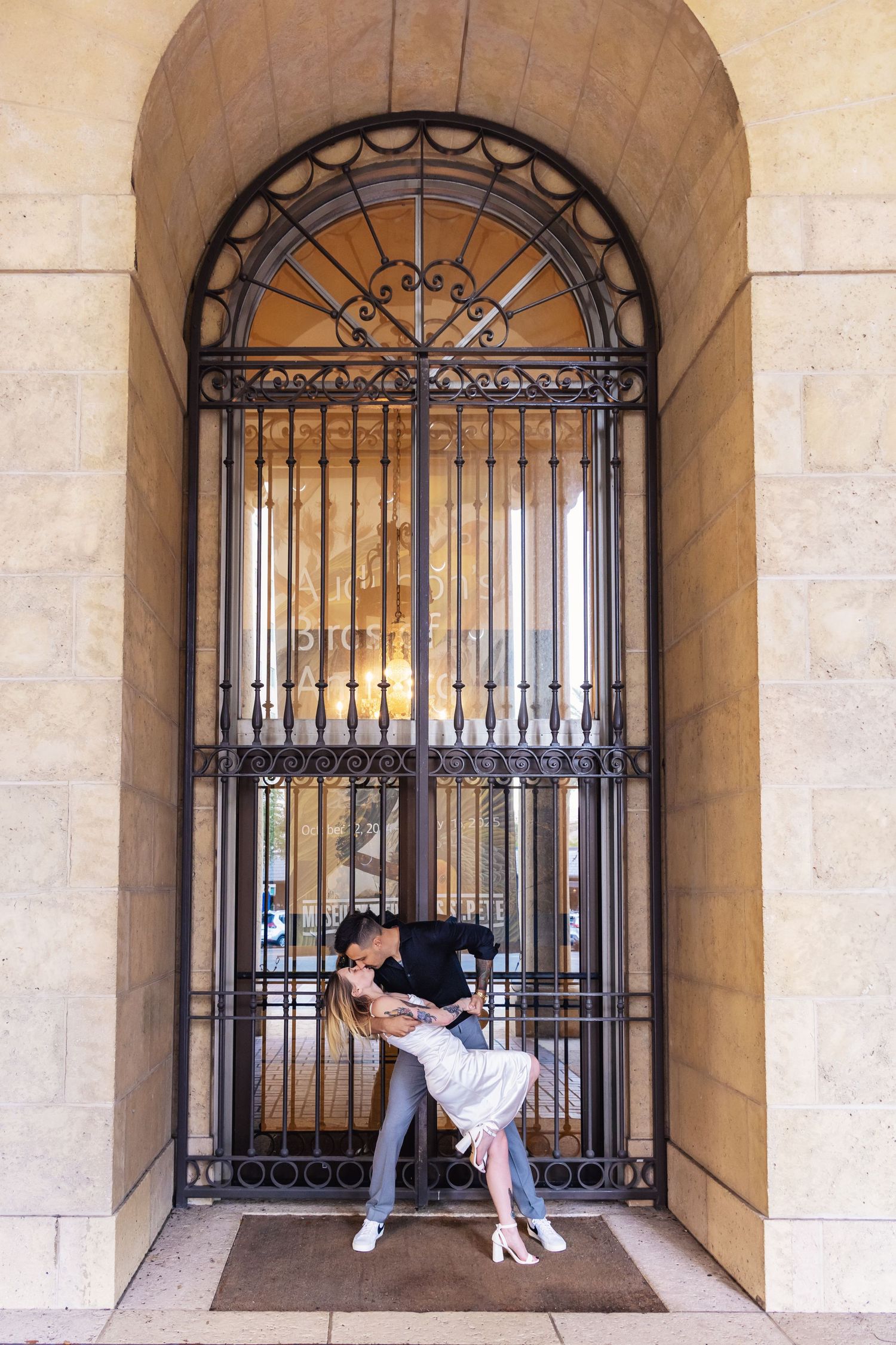 Romantic couple embracing under an ornate wrought iron archway with stone walls and classic architectural details.