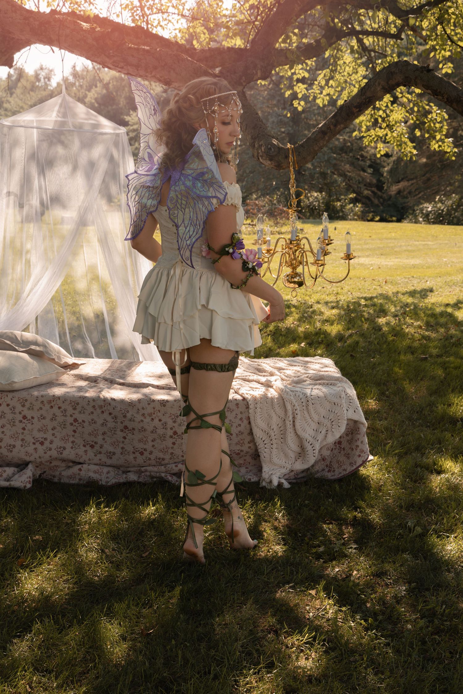 Woman in fairy costume with wings and green leg wraps poses outdoors beside a canopy bed in a sunlit garden.