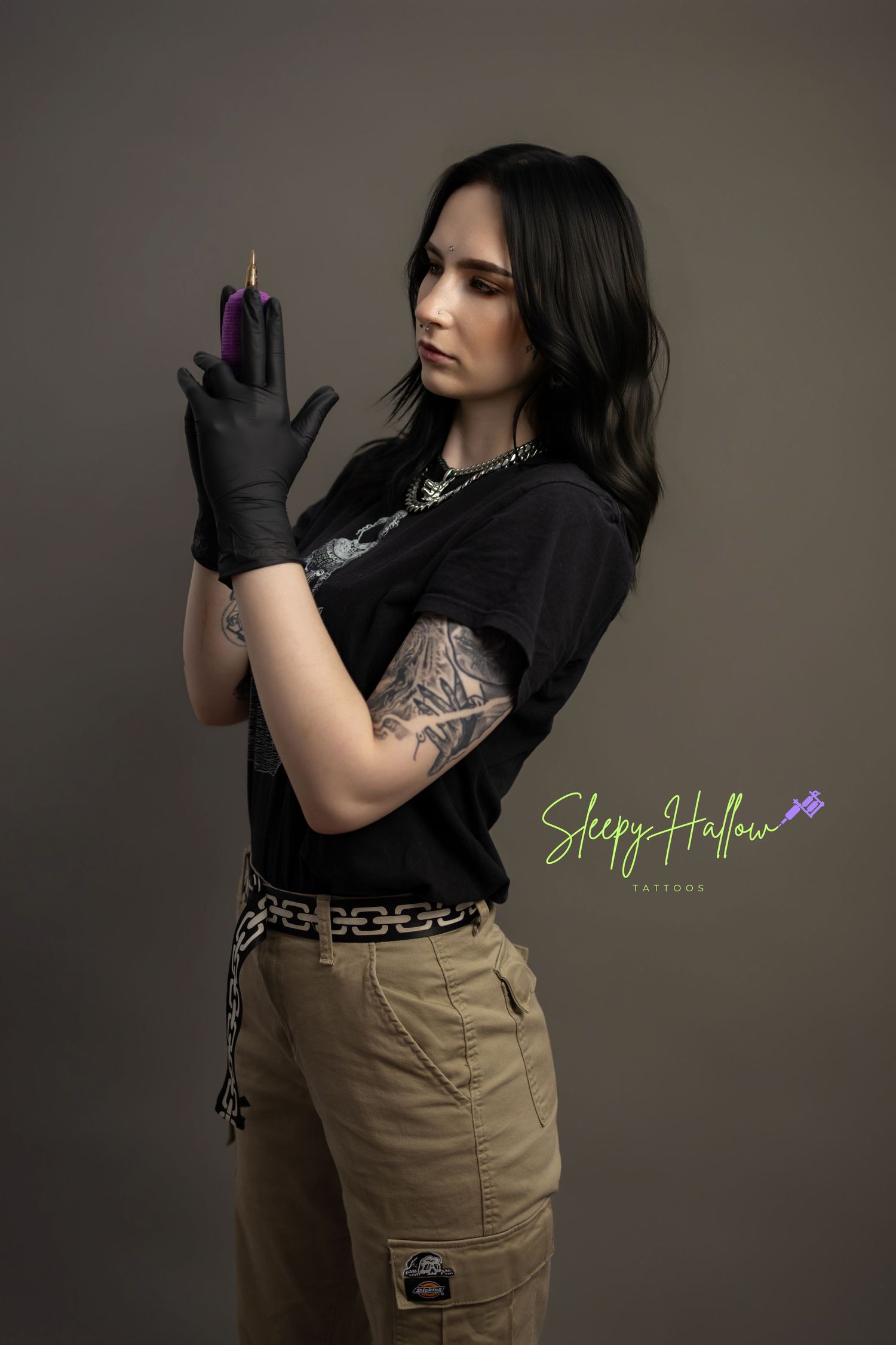 Tattooed woman in black gloves and t-shirt holds purple marker, promoting Sleepy Hollow Tattoos studio.