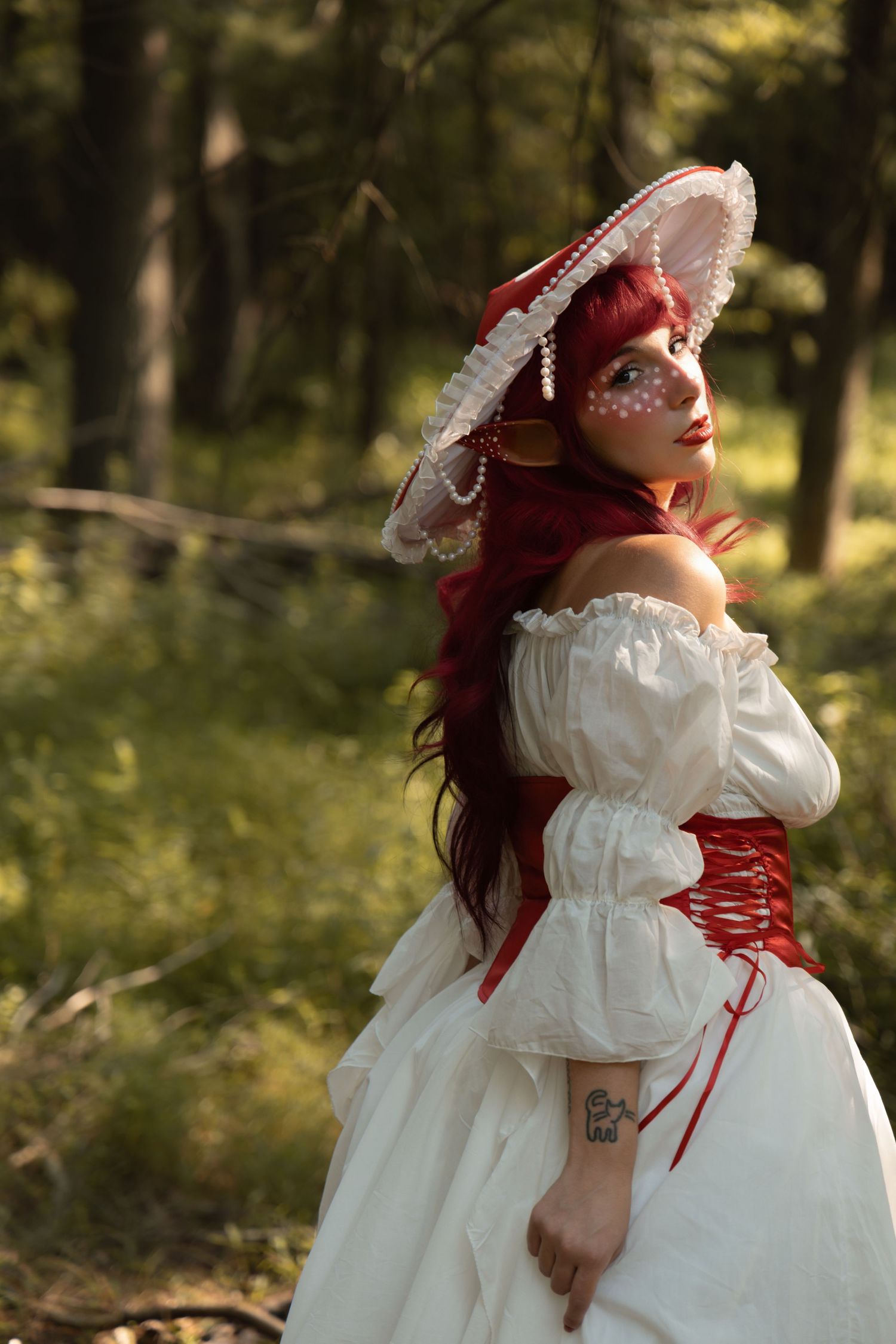 Woman in white off-shoulder dress, red corset, and lace hat with red hair poses in a forest setting.