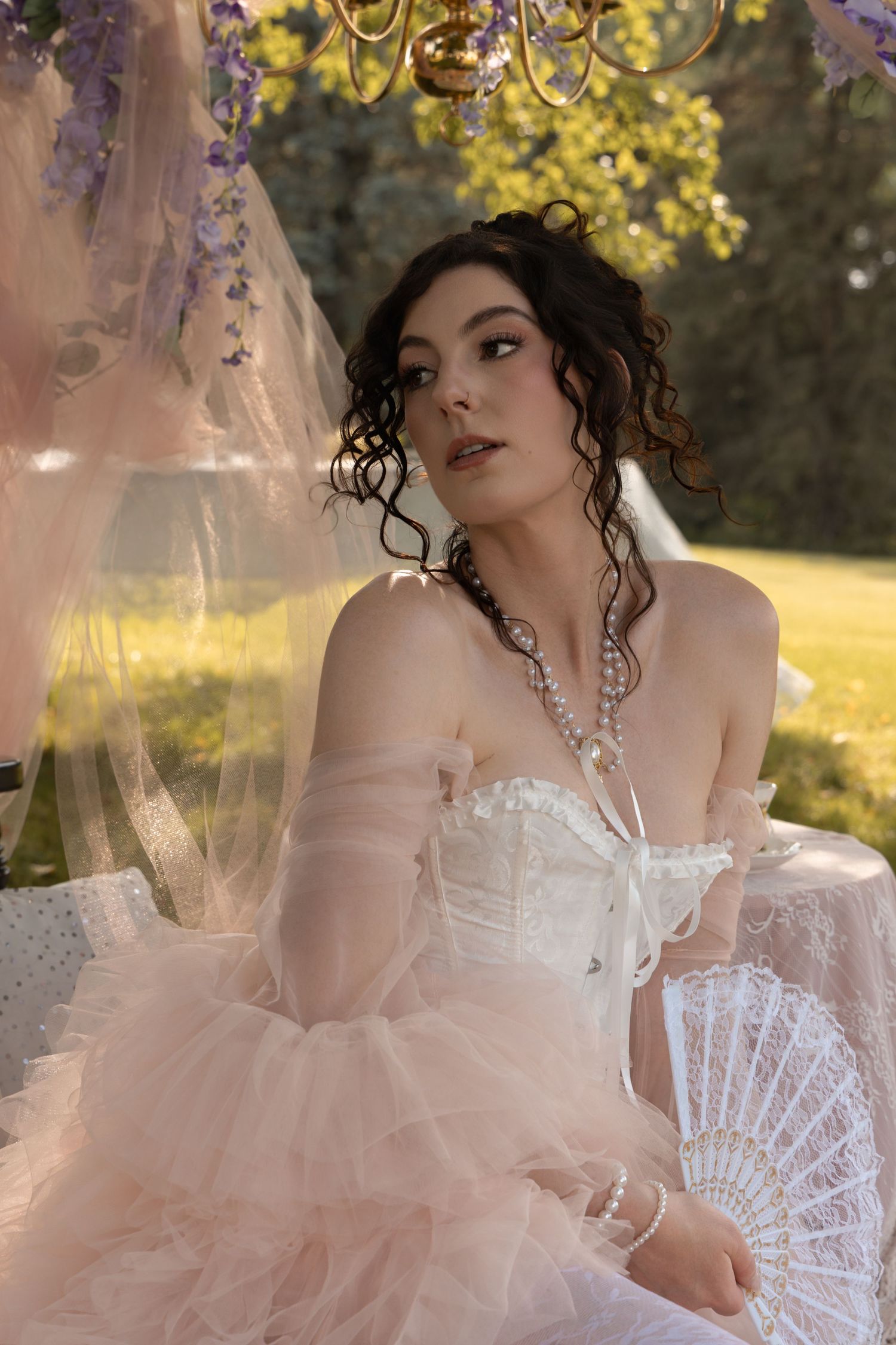 Young woman in pink tulle ballgown with corset and pearl necklace holds lace fan under wisteria chandelier outdoors.
