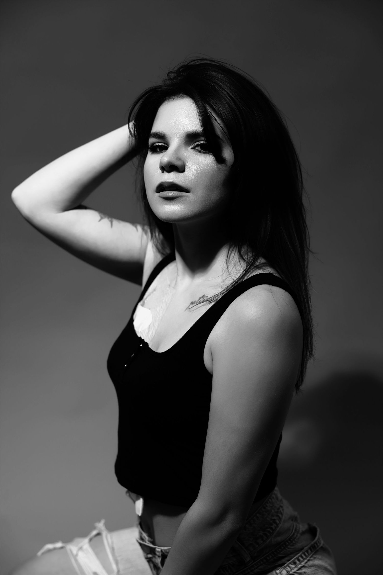 Black and white portrait of a young woman with long dark hair in a tank top, posing with hand behind her head.
