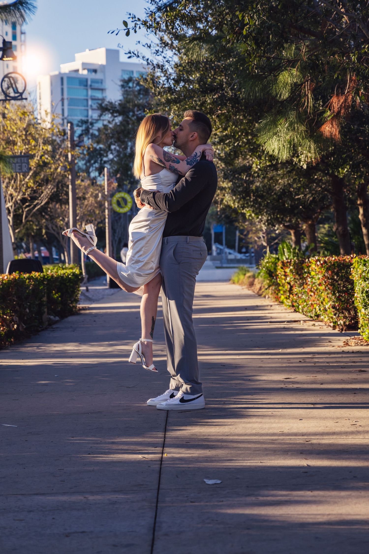 A couple dances together on a palm tree-lined pathway at sunset with urban buildings in the background.