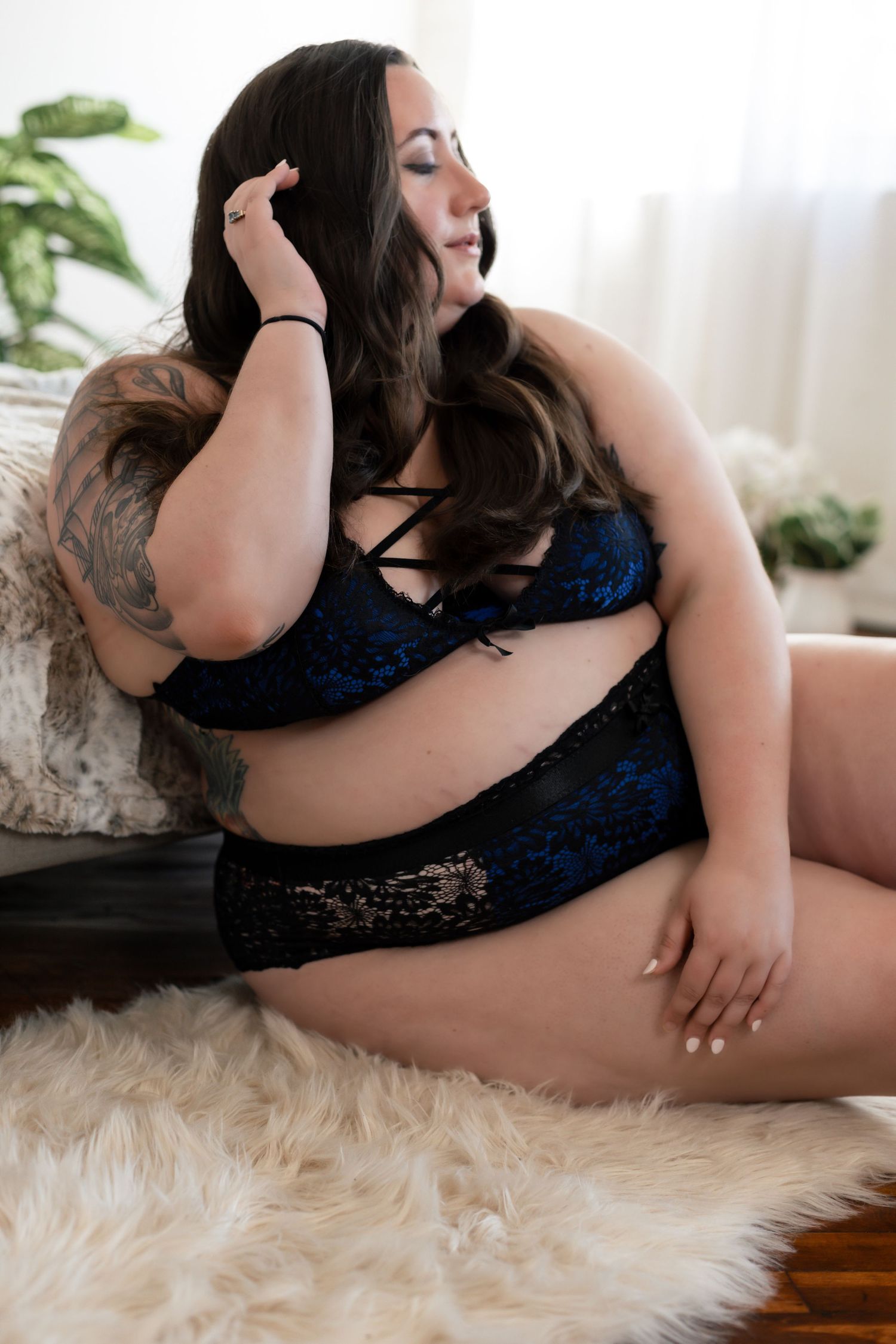 A person in blue lace lingerie poses on a white fur rug with houseplants visible in the background.