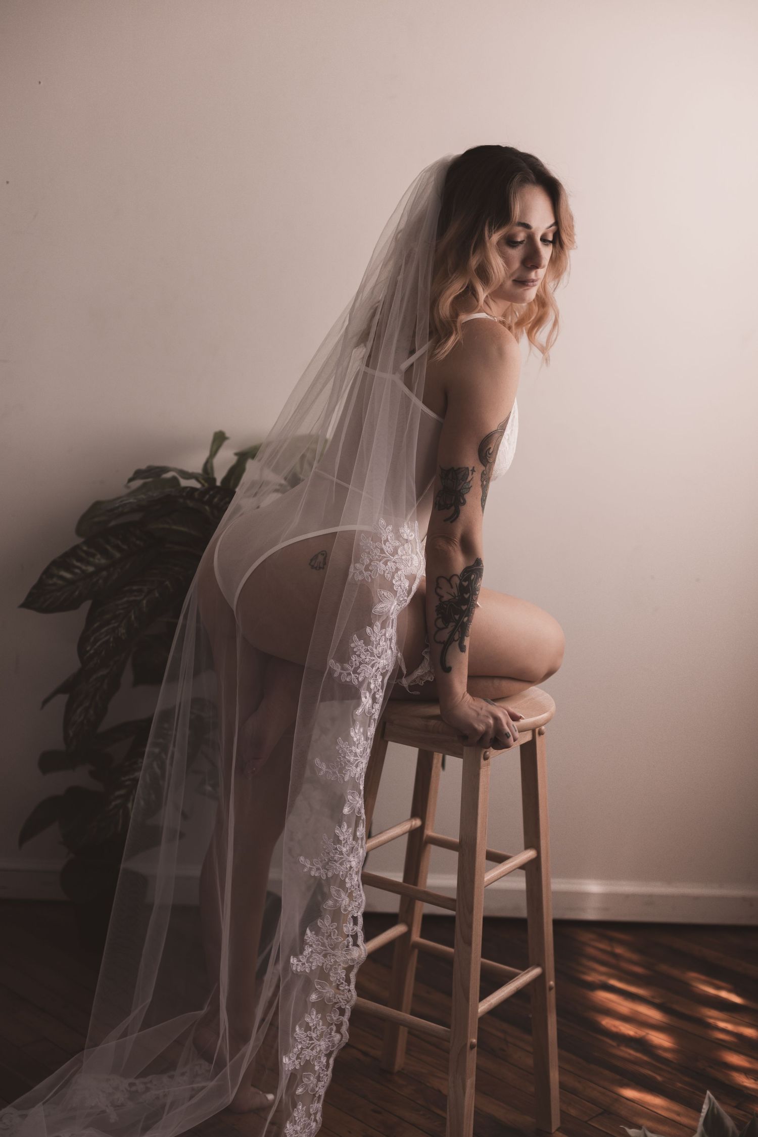 Woman in white lingerie and veil poses on wooden stool with potted plant in background.