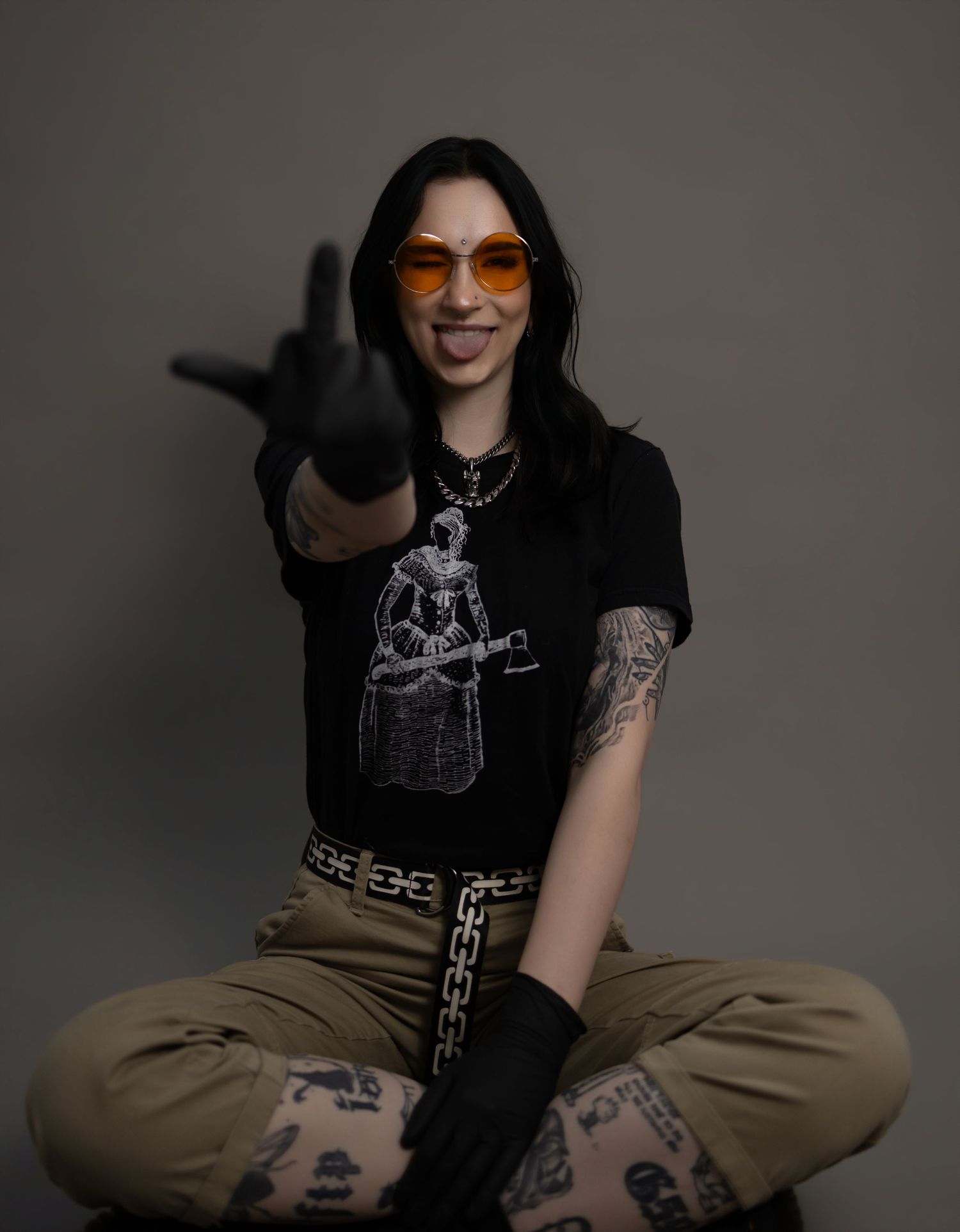Tattooed woman in orange sunglasses, black graphic tee, and khaki pants sits cross-legged making rock gesture.
