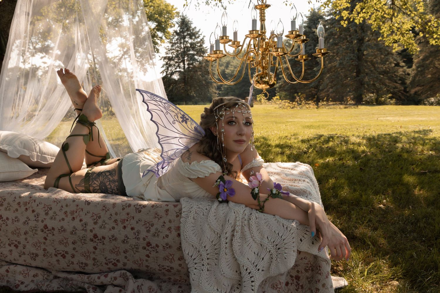 Woman dressed as a fairy with wings and floral crown resting on outdoor bed with chandelier in sunlit garden.