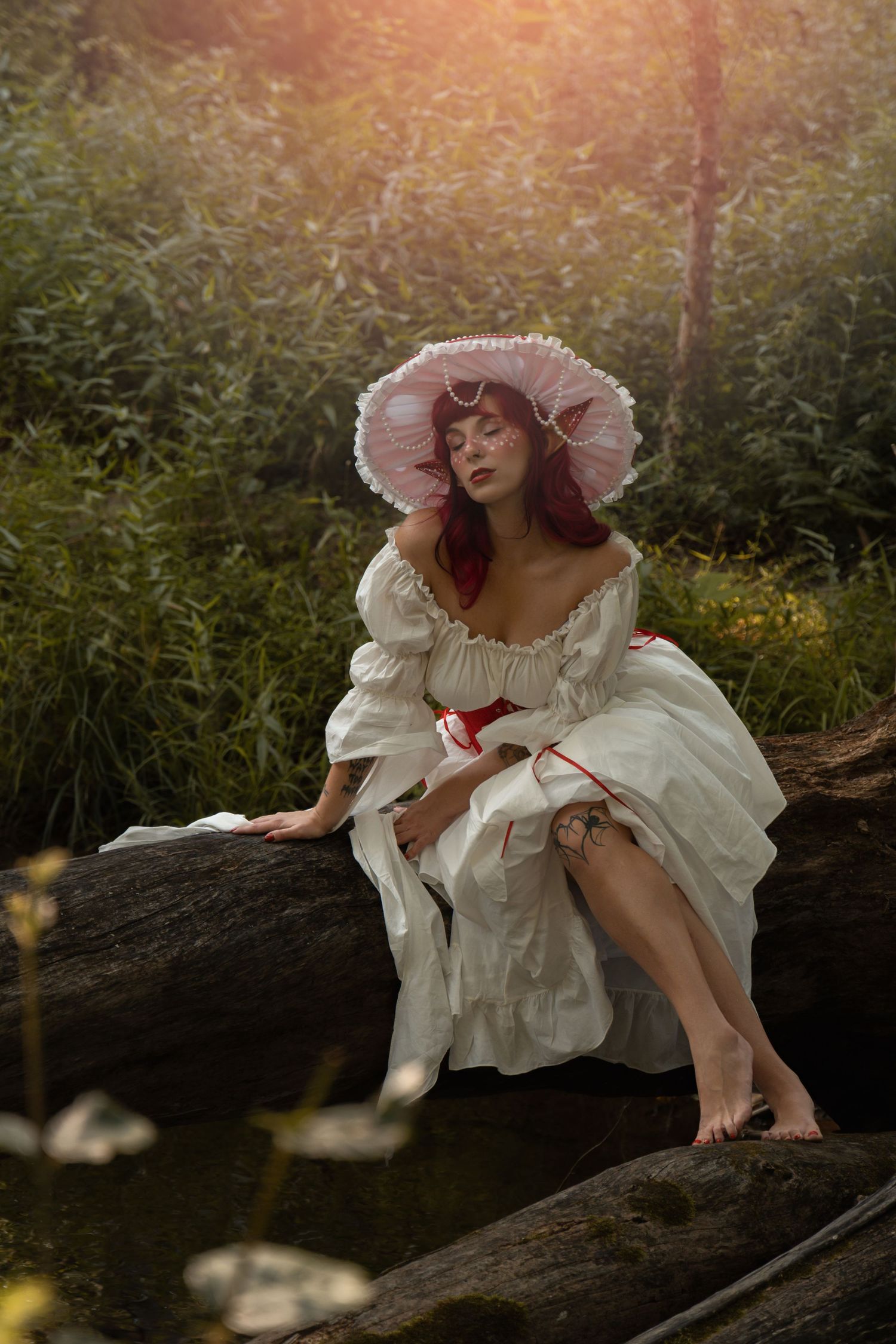 Woman in white off-shoulder dress and pink bonnet sits barefoot on a log in a lush green forest setting.