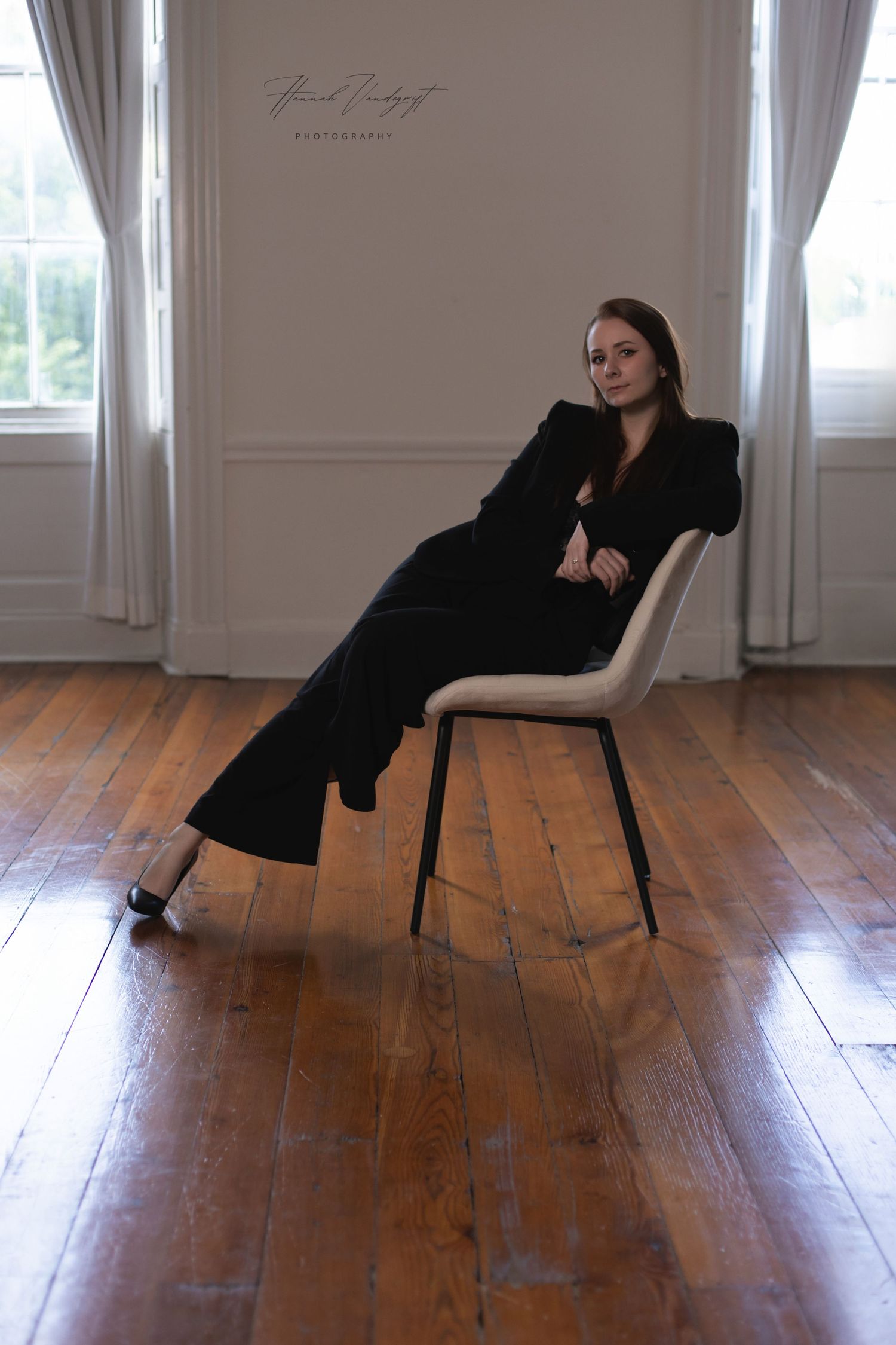 Professional fashion portrait in moody lighting featuring black pantsuit and white accent chair on hardwood floor.