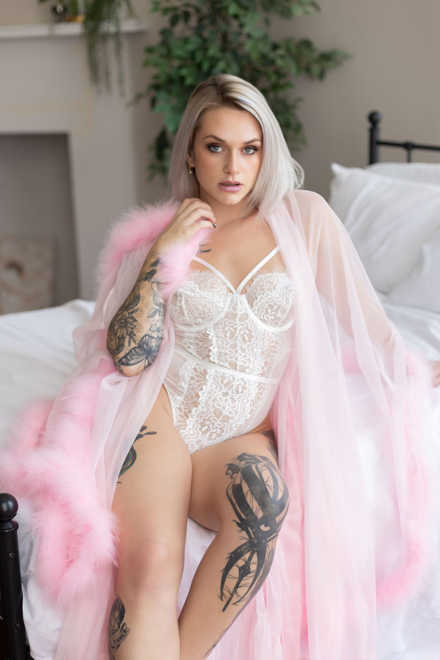 Blonde woman with tattoos wearing white lace lingerie and pink feather robe sitting on a bed.