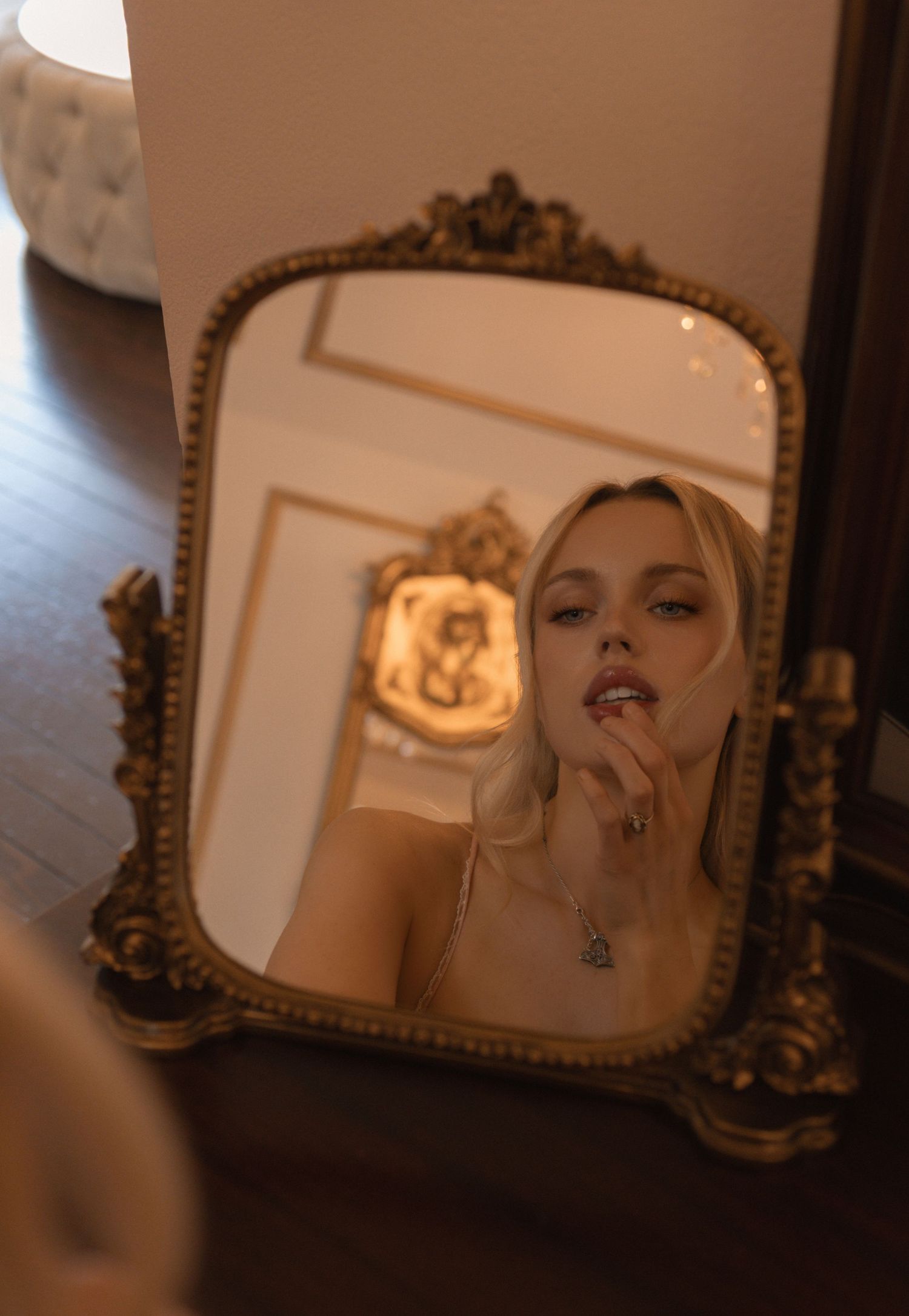 A blonde woman gazes into an ornate gold-framed mirror, touching her lips while wearing a delicate necklace.