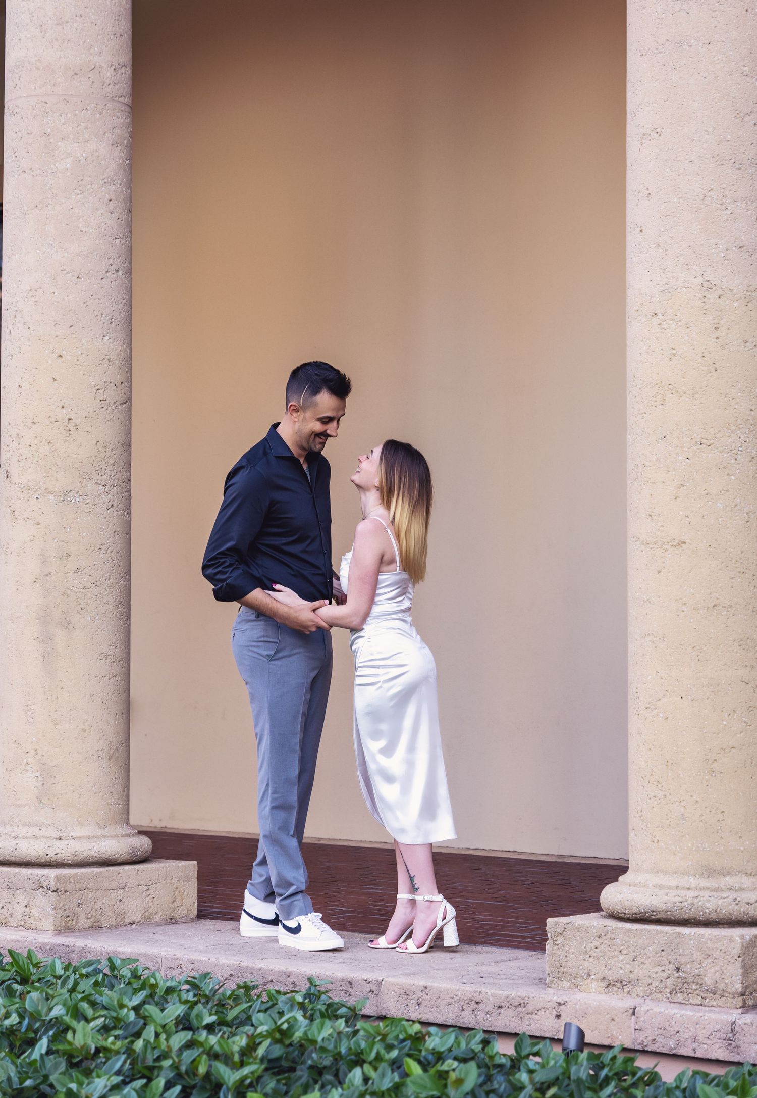 A couple shares a romantic moment between elegant stone columns in formal attire at dusk.