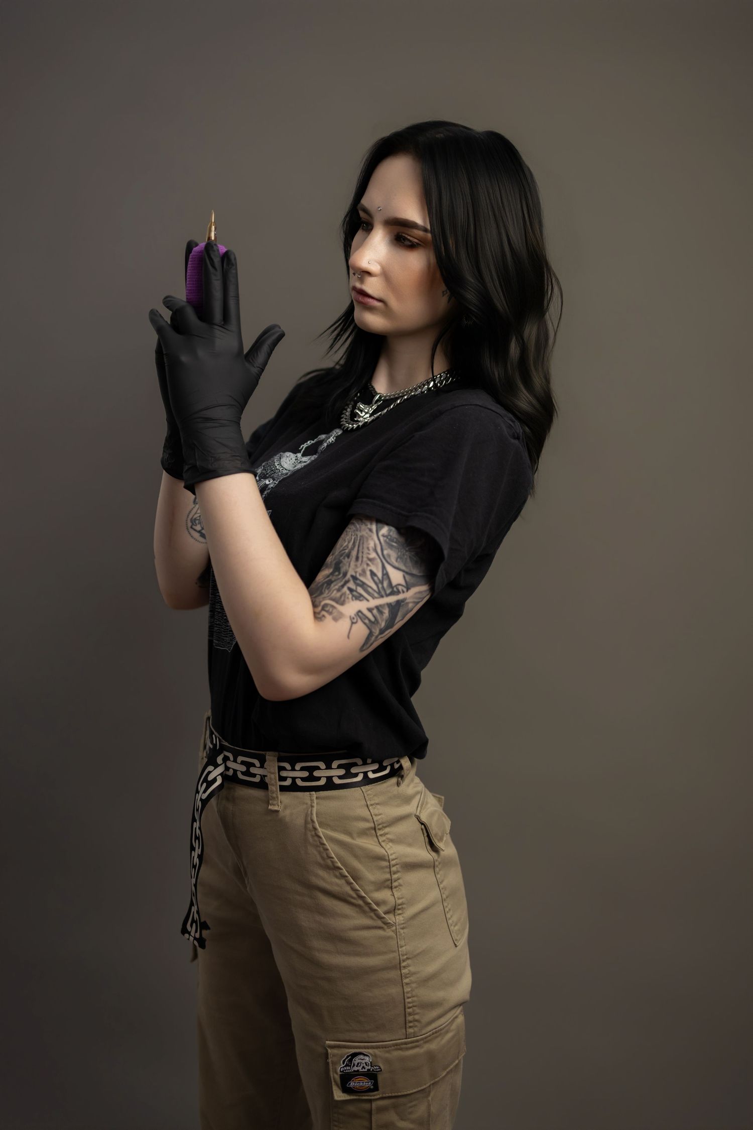 Female tattoo artist in black gloves examining a purple tattoo marker, showcasing sleeve tattoos in a studio setting.
