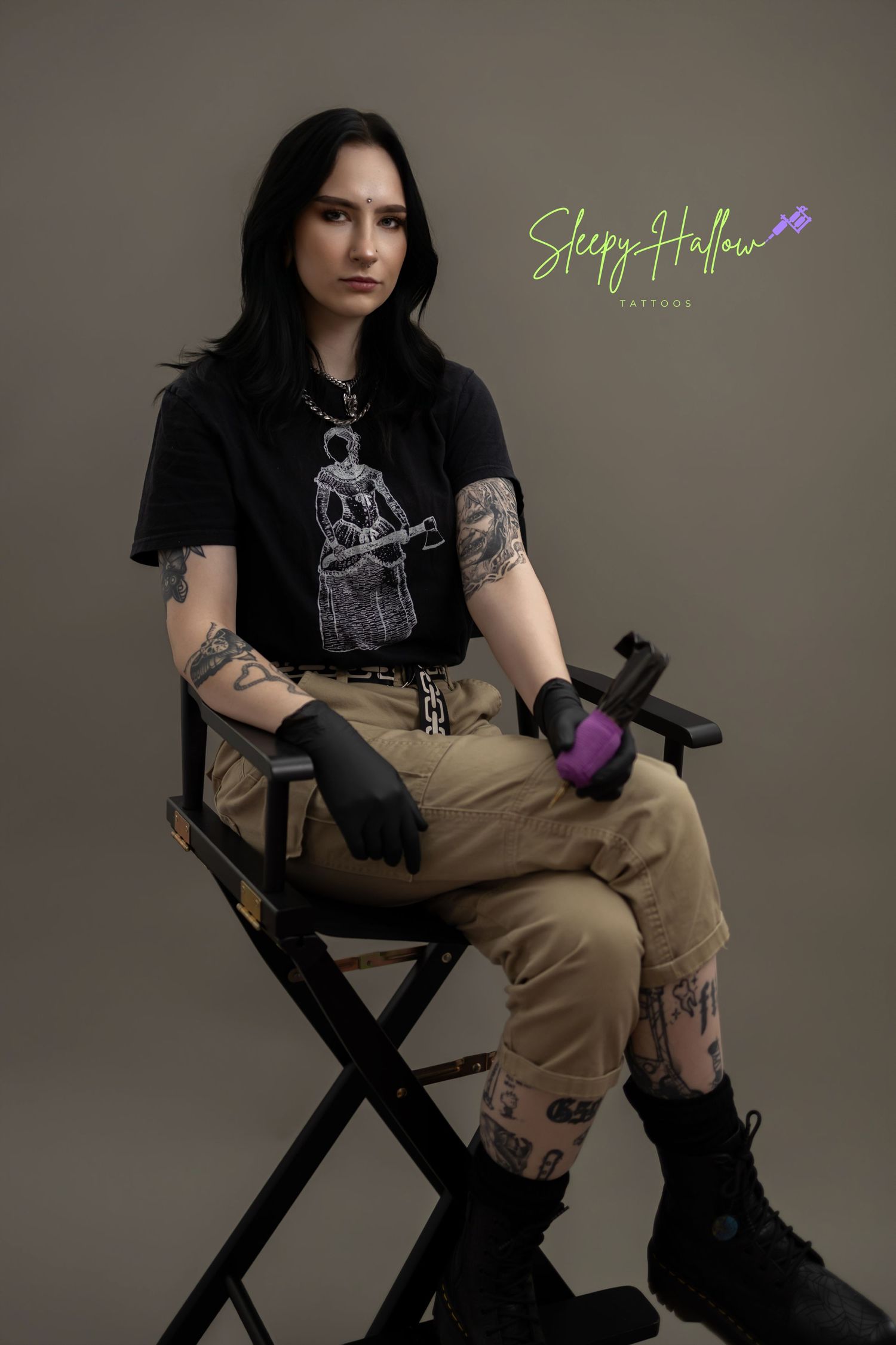 Tattooed tattoo artist sitting in director's chair wearing black gloves and tee, khaki pants, boots, Sleepy Hollow Tattoos.