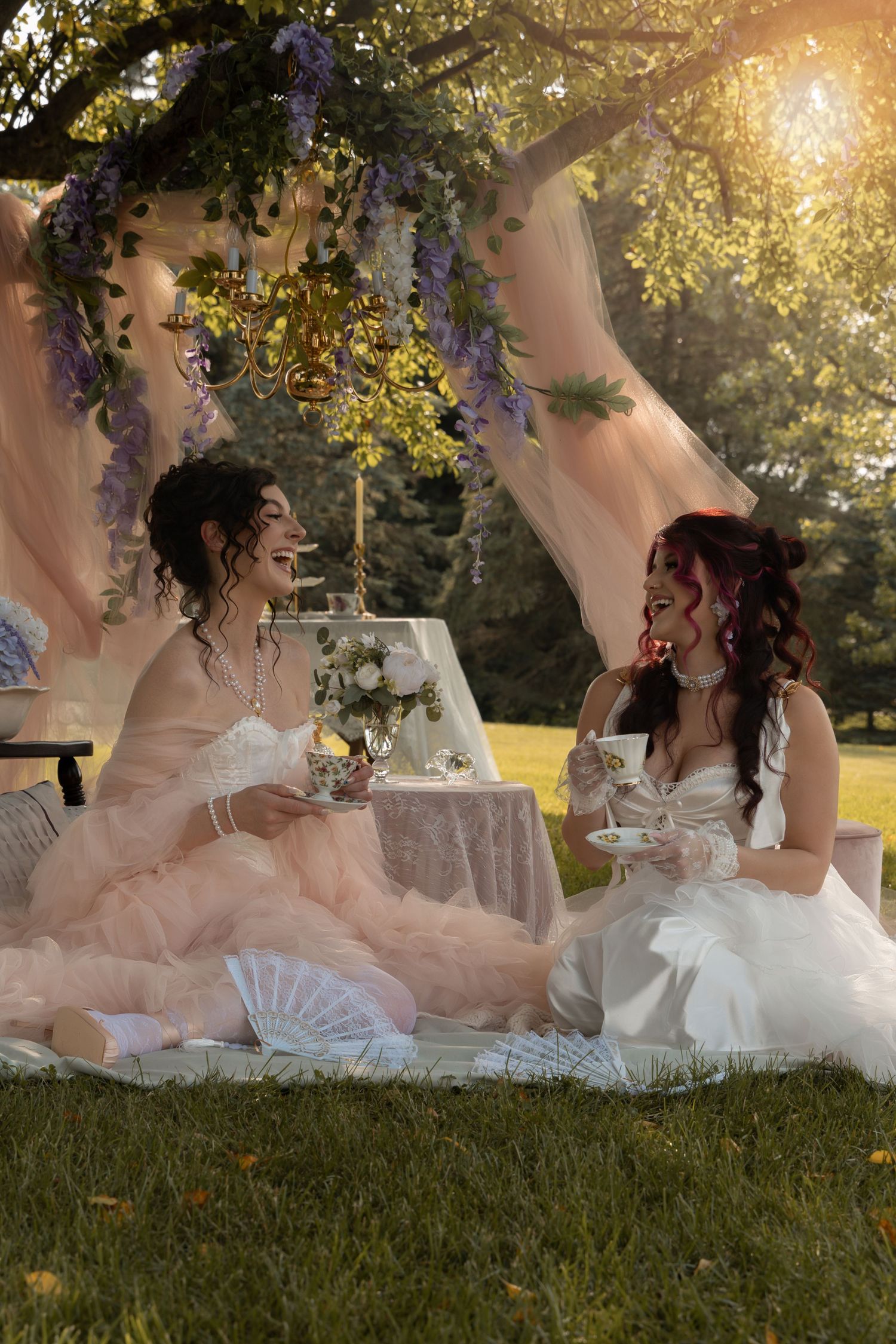 Two brides in elegant white gowns enjoy an outdoor tea party under wisteria-draped canopy in golden sunlight.
