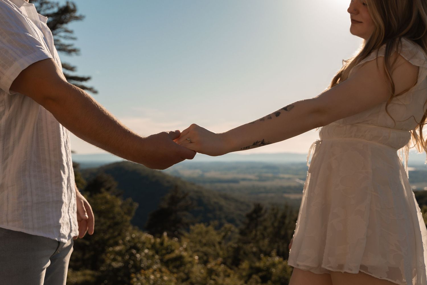 Couple holding hands on a scenic mountain overlook with lush green trees and blue sky in the background.
