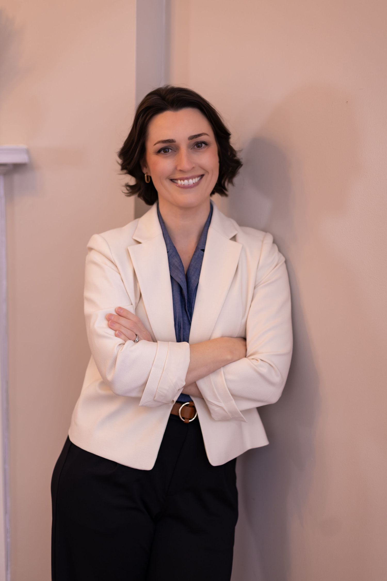 Professional woman in white blazer and navy top stands with crossed arms in office.