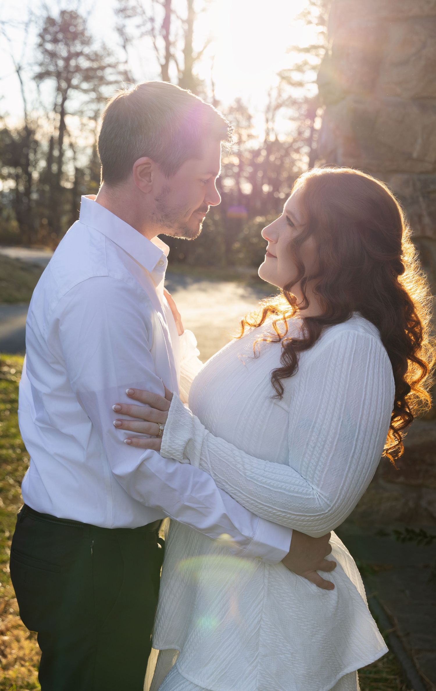 A couple embraces in warm sunlight while wearing white sweaters during a romantic outdoor photo session.