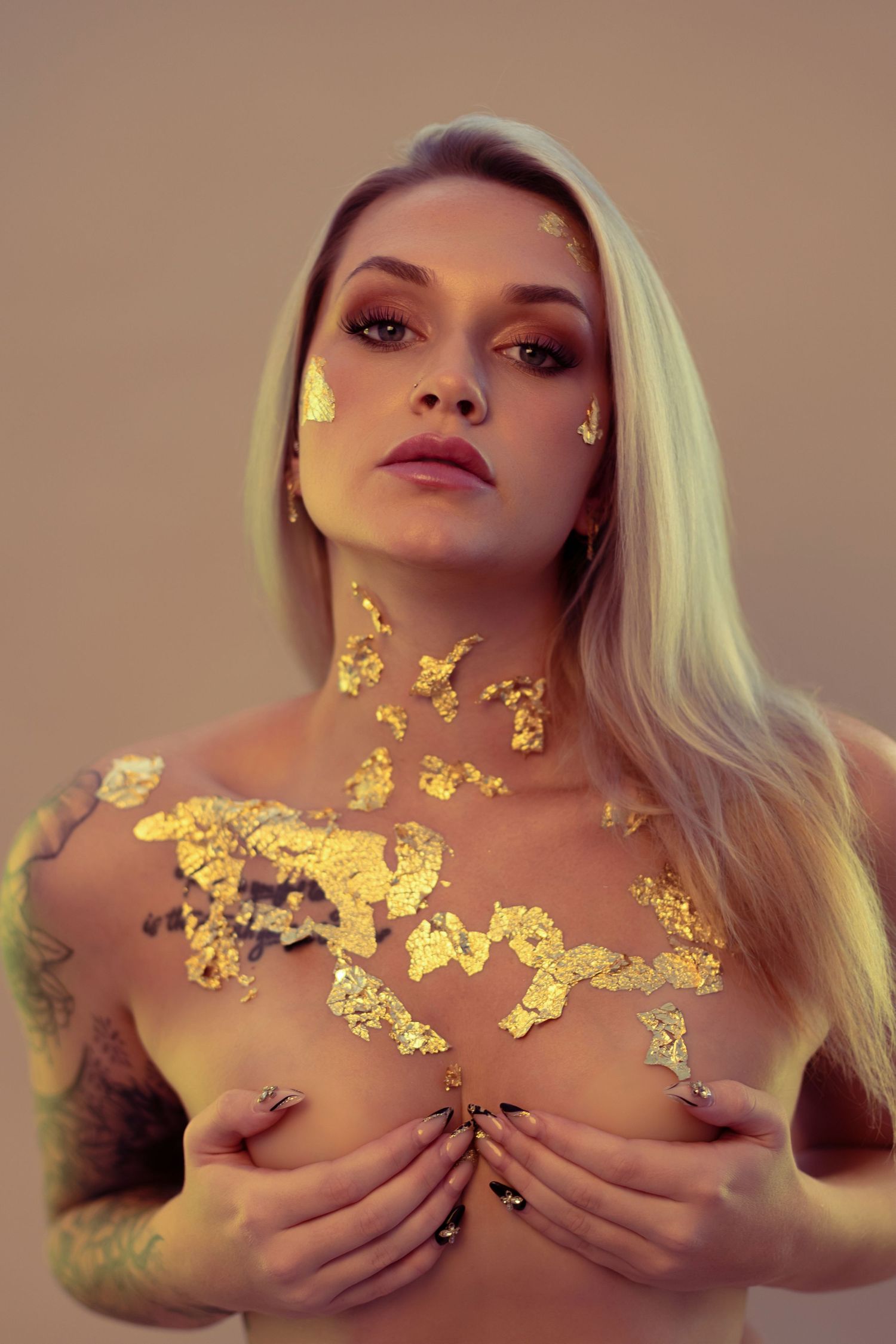 Blonde woman with gold leaf flakes on her face, neck, and chest, posing confidently in a studio setting.