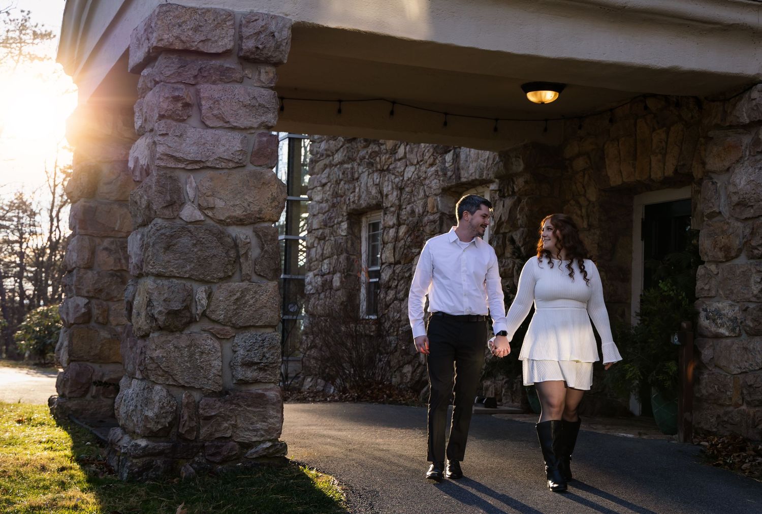 A couple walks hand in hand along a stone building exterior at sunset, with warm sunlight streaming through the pillars.