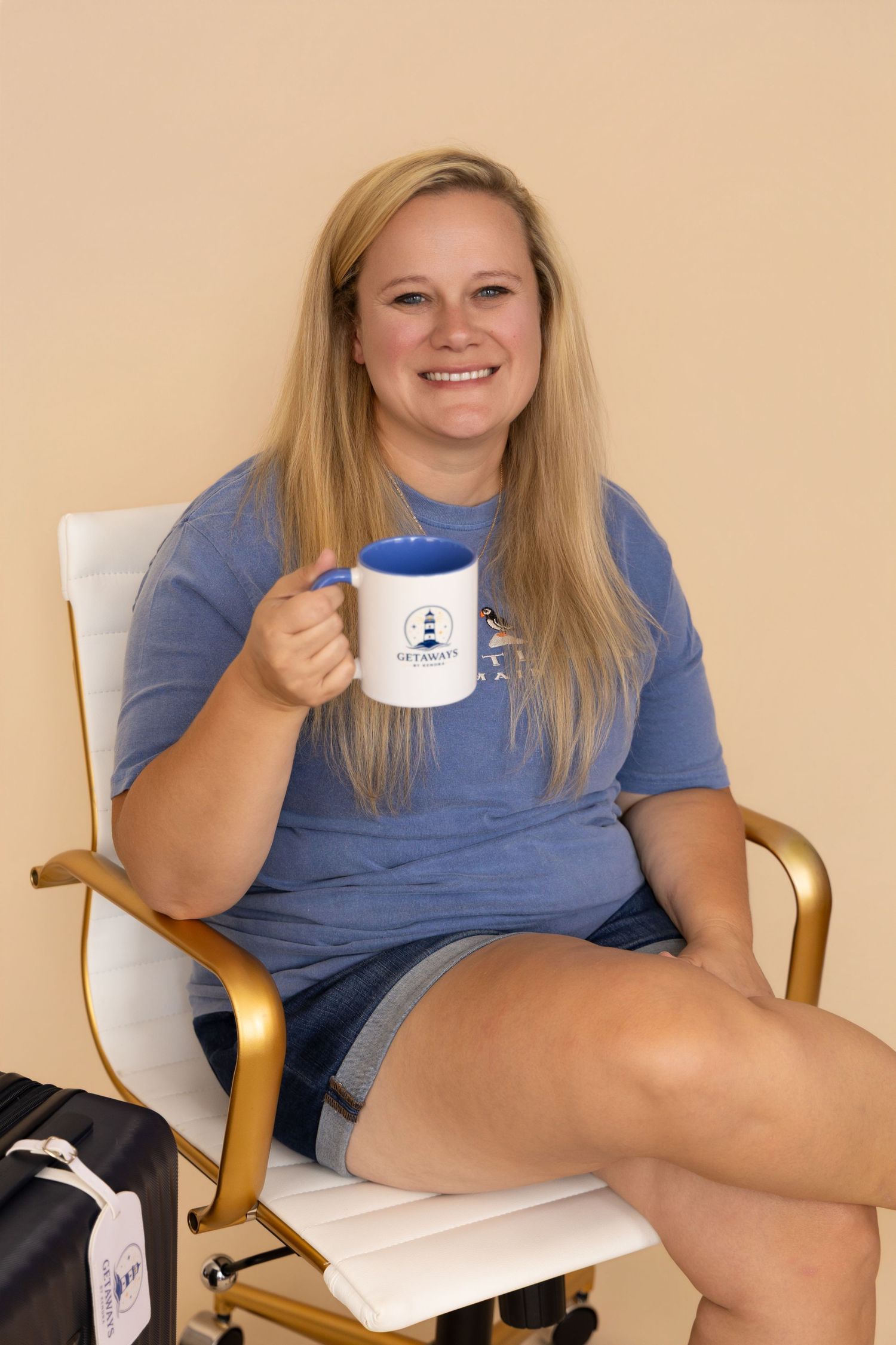 Smiling blonde woman sitting in gold chair holding blue and white lighthouse mug, wearing blue tee and denim shorts.