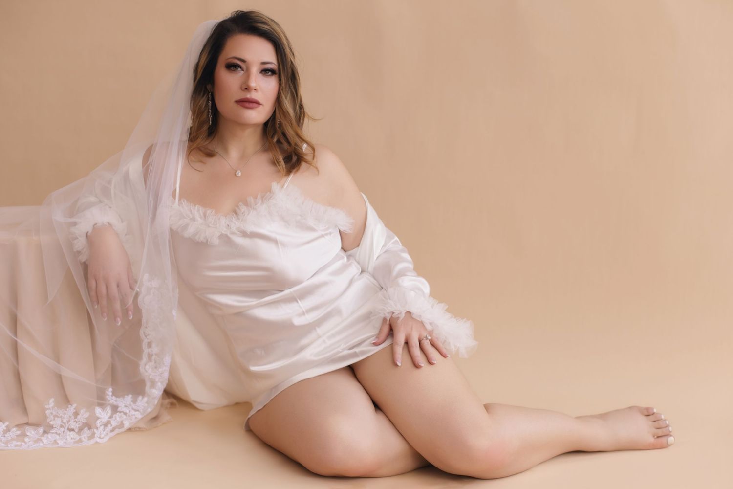 Figure in white fabric draping poses sensually against beige backdrop in intimate boudoir setting.