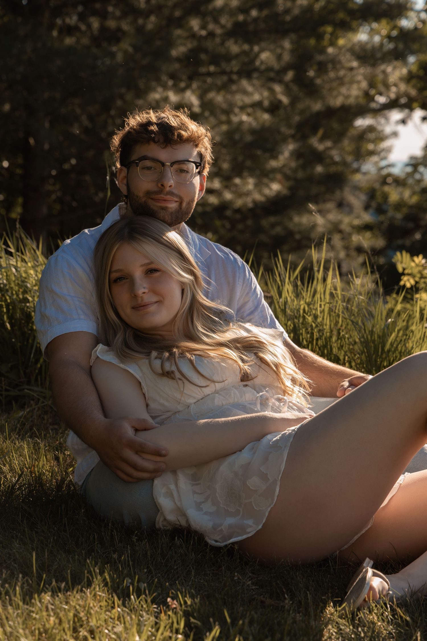 Couple sitting in grass at golden hour, woman leaning back on man wearing glasses, surrounded by lush greenery.