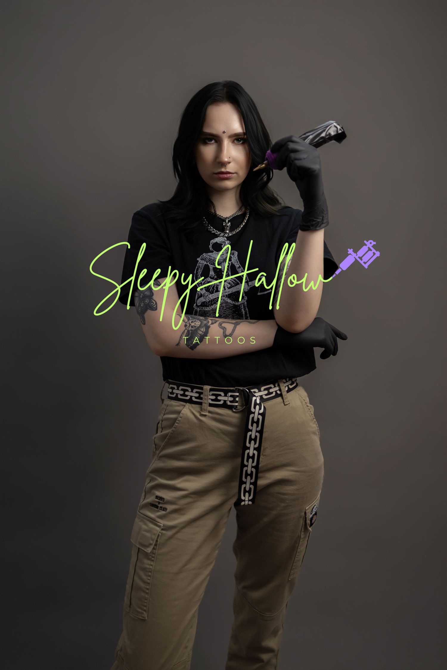 Tattoo artist holding a tattoo machine with crossed arms, wearing black gloves and cargo pants, Sleepy Hallow Tattoos branding.