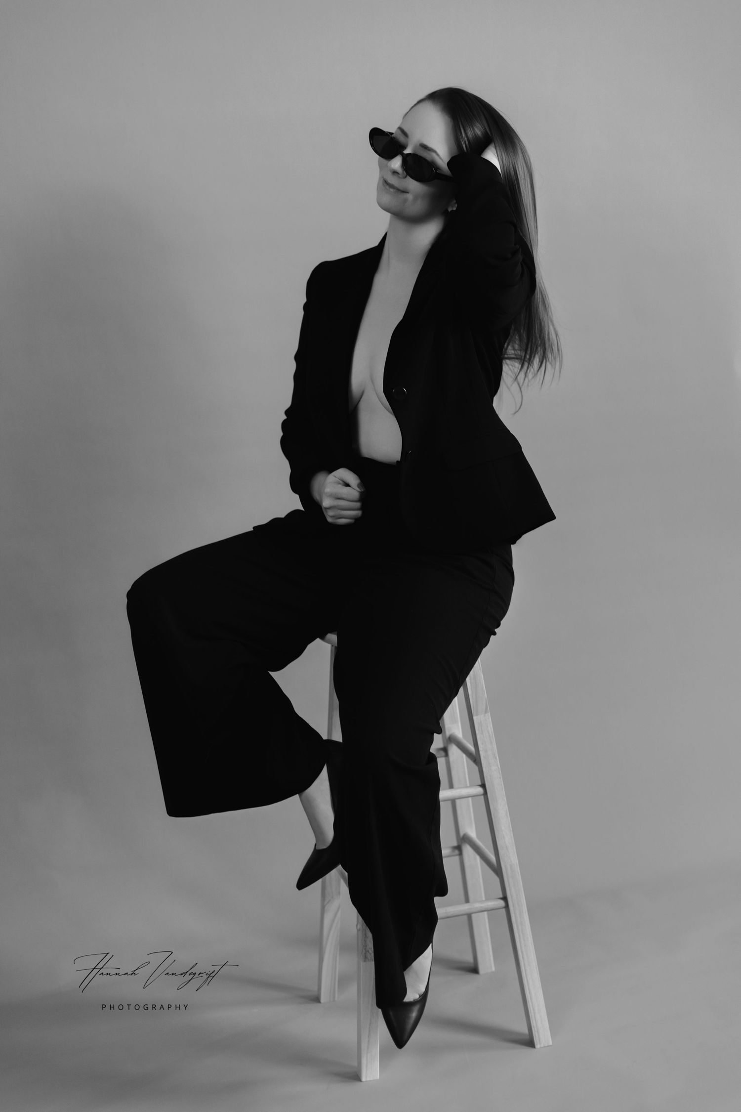 Fashion portrait of model in black blazer and sunglasses sitting on white stool.