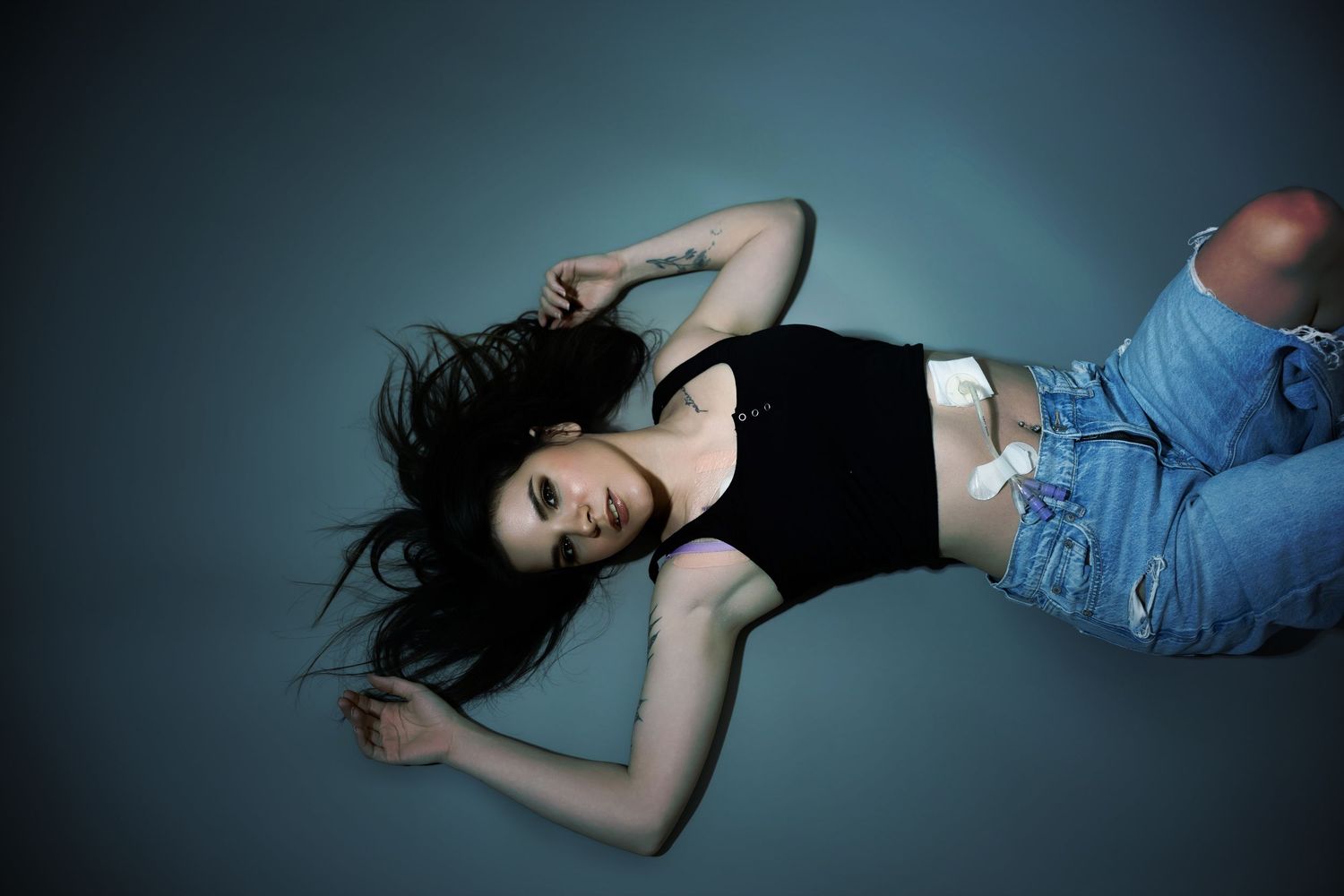 Young woman with dark hair lying on floor wearing black crop top and ripped blue jeans in dramatic studio lighting.