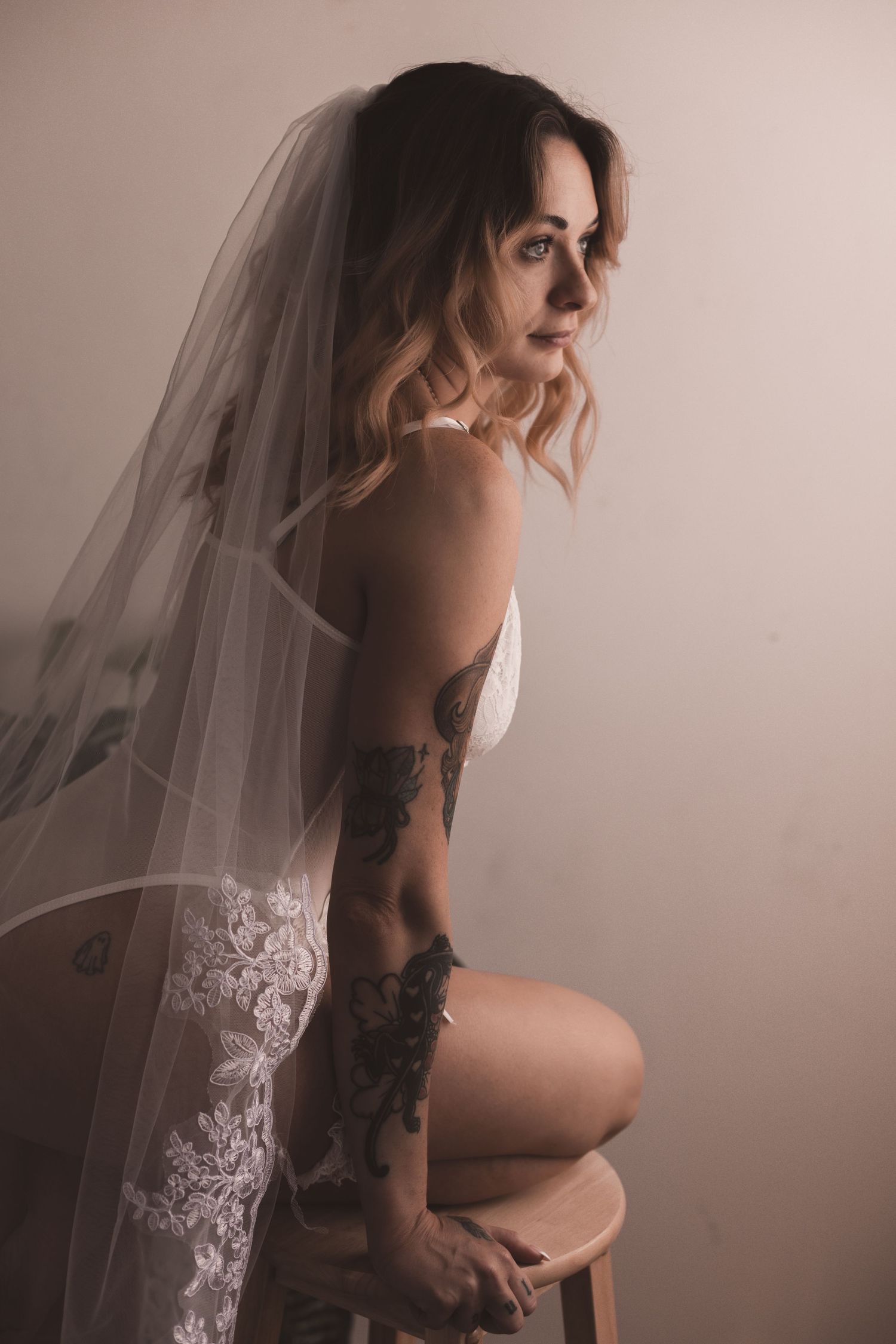 Woman in white lingerie poses on stool with flowing lace veil.