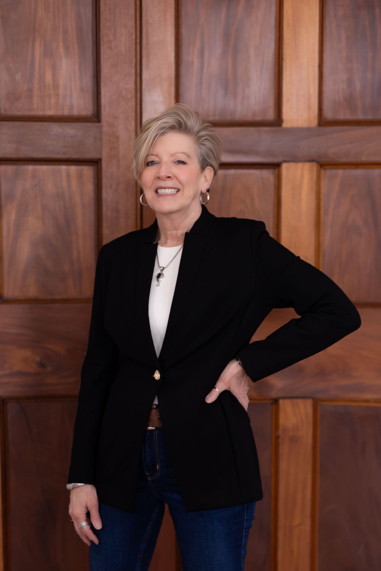 Business professional in black blazer and jeans poses confidently against wooden paneled wall.