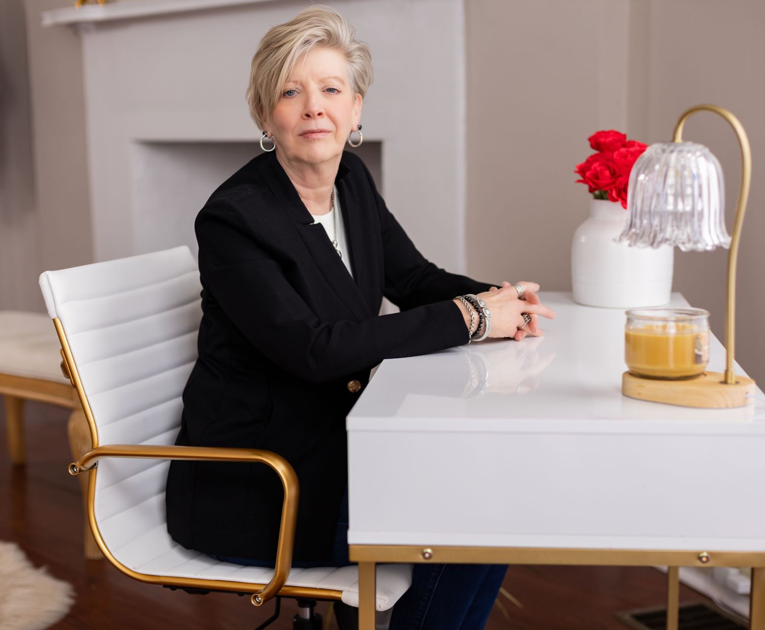 Professional woman in black blazer sits at modern white desk with table lamp.
