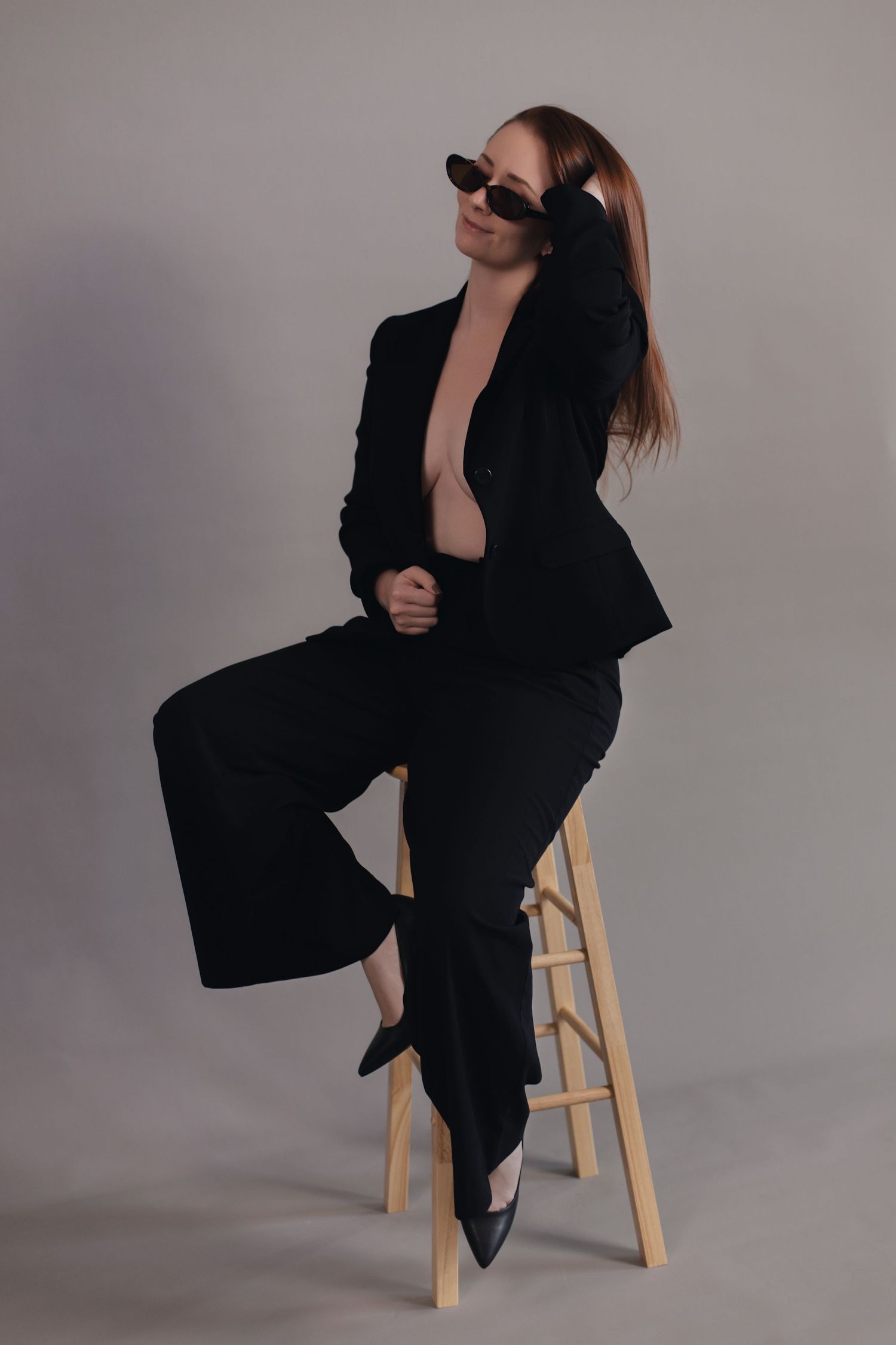 Artistic fashion poses on wooden stool against grey backdrop wearing all black ensemble.