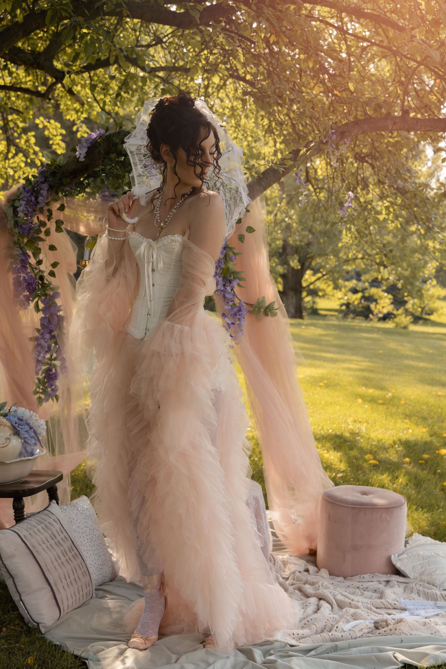 Woman in pink ruffled gown and lace veil stands under flowering tree at outdoor garden wedding picnic.